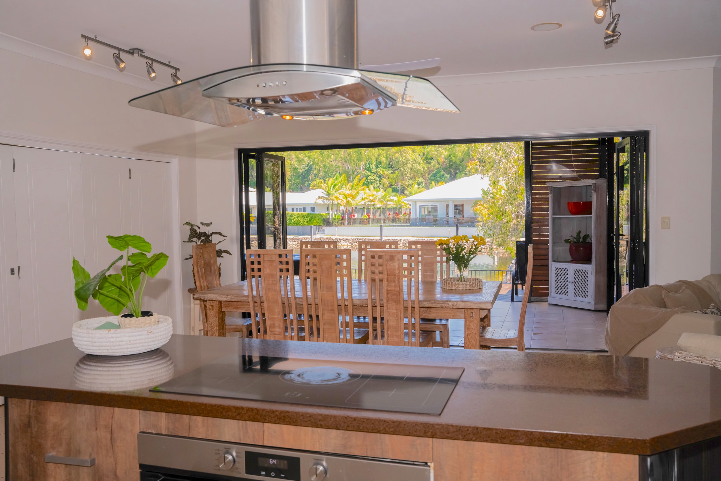 Modern kitchen and dining area with island countertop, induction stove, wooden dining table and chairs, large open sliding glass doors leading to an outdoor area, potted plants, and ceiling lights. Outside view shows greenery and a distant house.