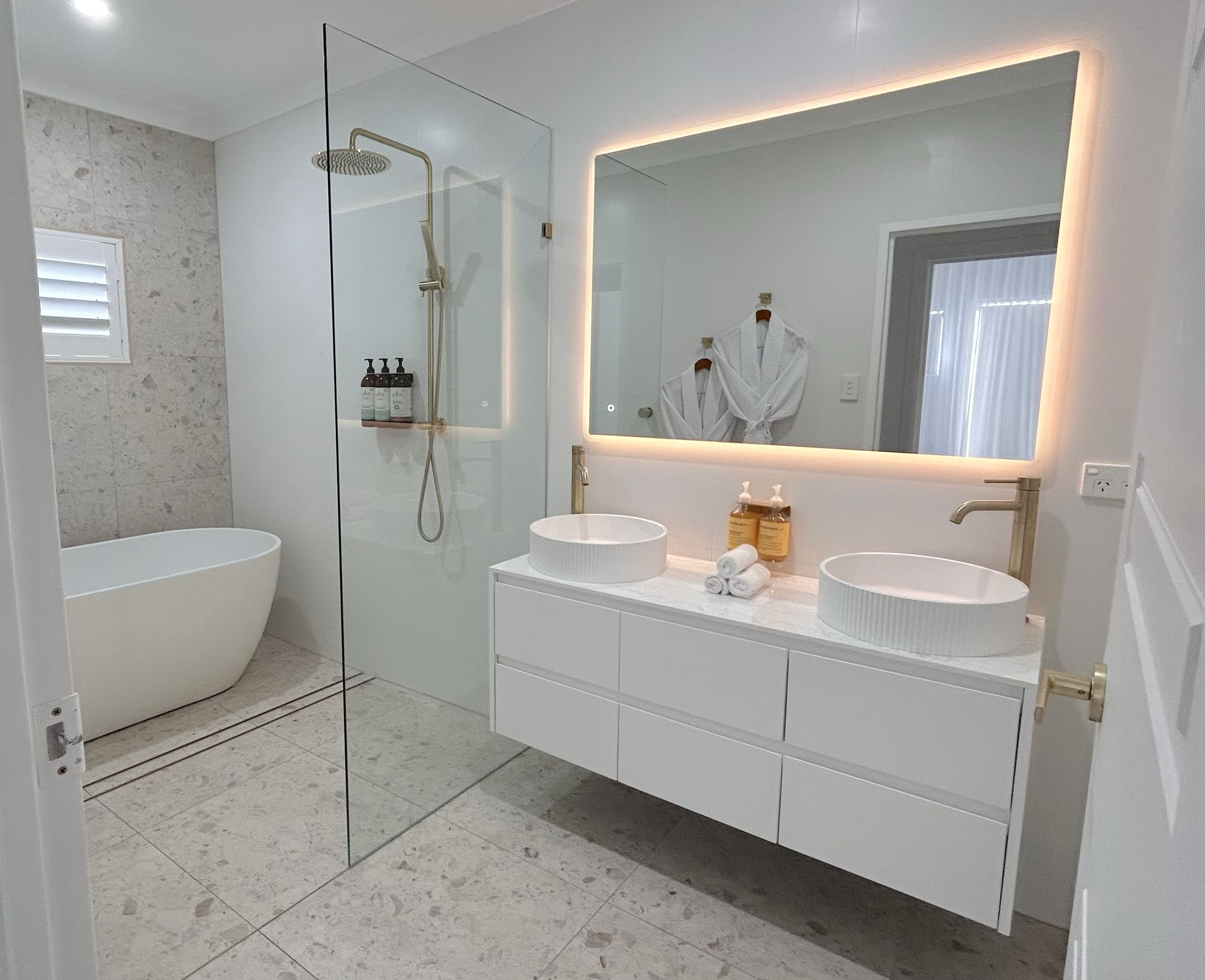 Modern bathroom with a freestanding bathtub, glass-enclosed shower, double vanity with vessel sinks, illuminated mirror, and bathrobes hanging on the wall.