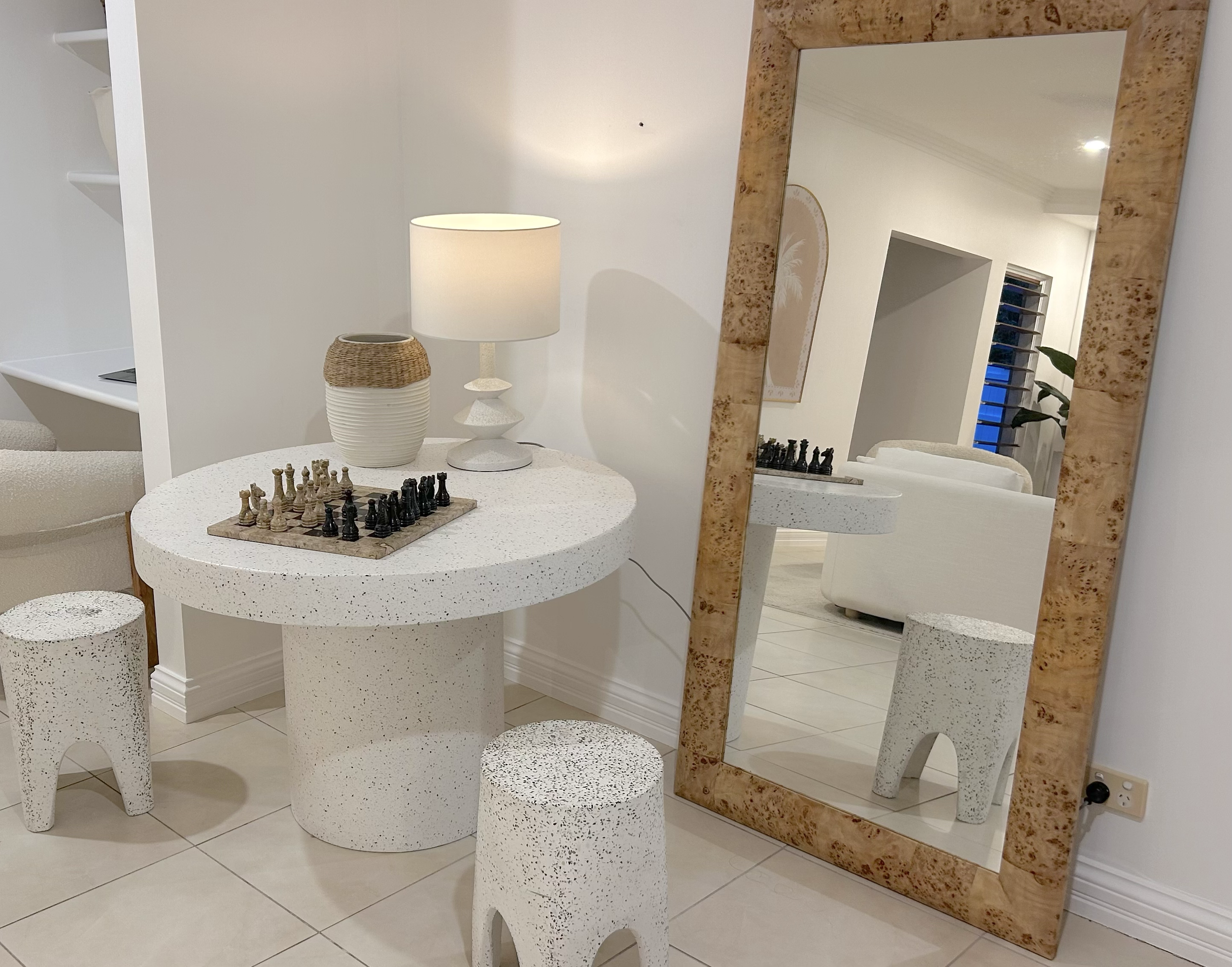 Modern living room with a white speckled round table featuring a chess set, two matching stools, a large wooden-framed mirror, and a decorative lamp with a woven detail.