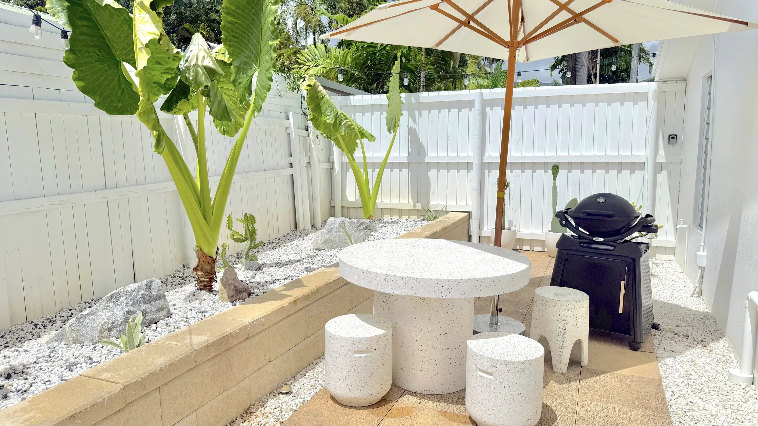 A backyard patio with white gravel, two large potted tropical plants, a white circular table with matching stools, a black grill, a large beige patio umbrella, white fencing, and string lights overhead.