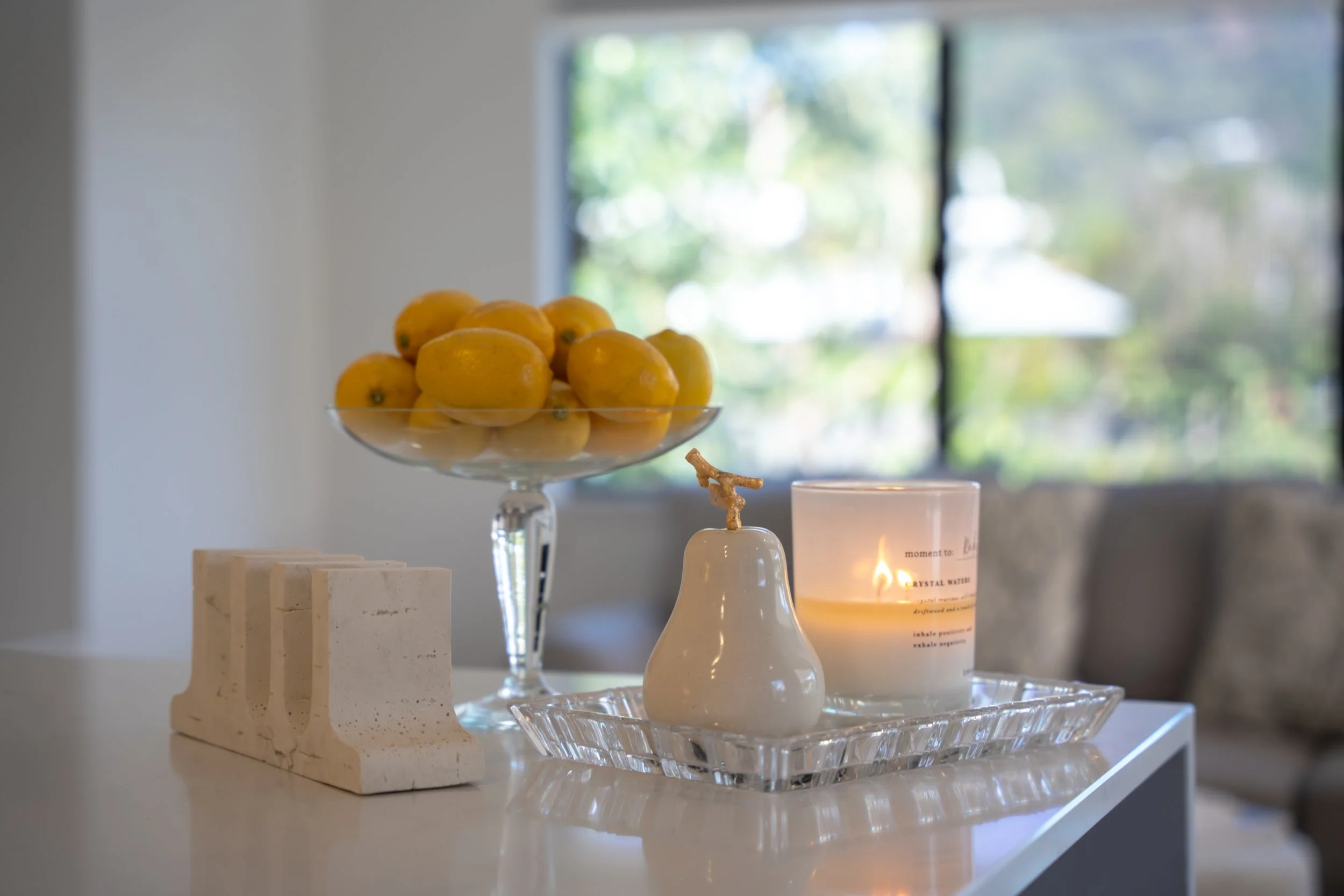 A glass bowl filled with yellow lemons on a transparent glass tray, next to a white candle and a carved soap stone, with a window in the background.
