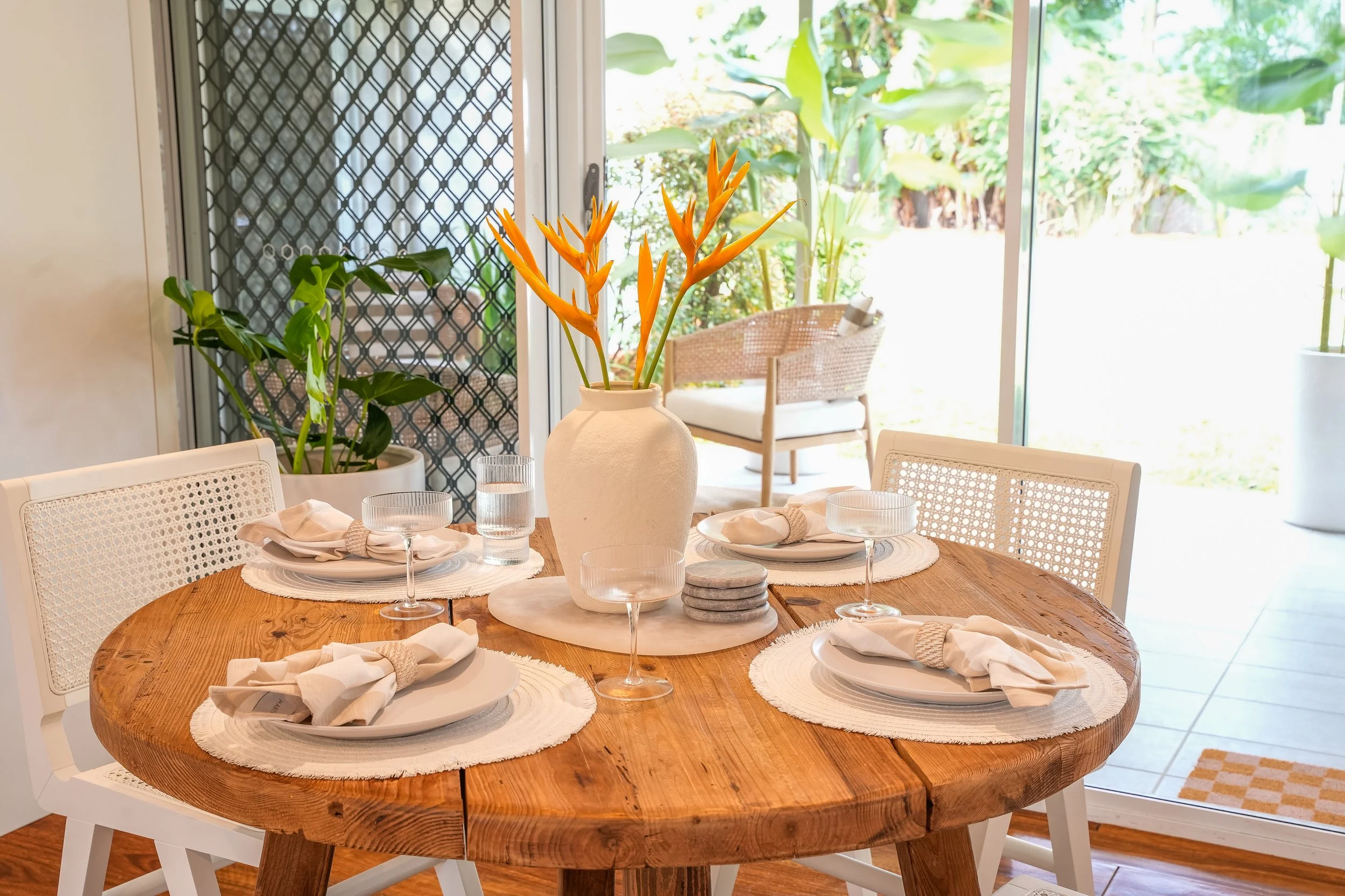 A round wooden dining table set with white plates, beige cloth napkins, and clear drinking glasses. In the center is a large white vase with orange tropical flowers. There is a large green plant in a white pot on the left side and a sliding glass door with a screen door behind the table, leading to a lush outdoor garden area with green foliage.