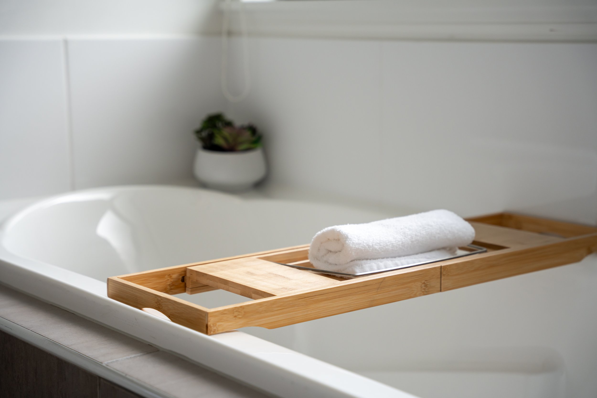 A white towel rolled up on a wooden tray placed on a white bathroom counter, with a bathtub and a small potted plant in the background.