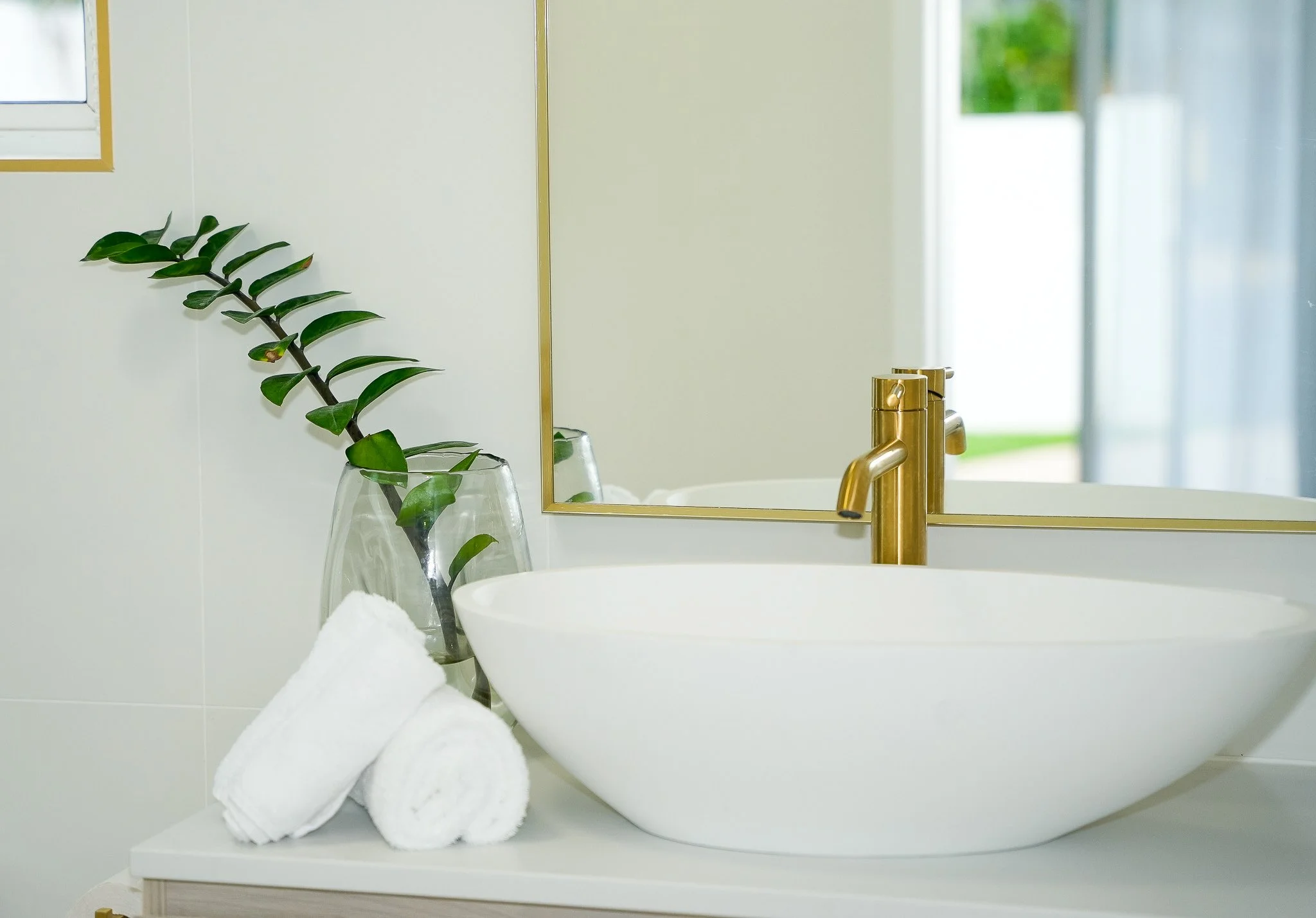 A bathroom counter with a white vessel sink, a gold faucet, a glass vase with a green plant, and two rolled white towels, with a mirror and window in the background.
