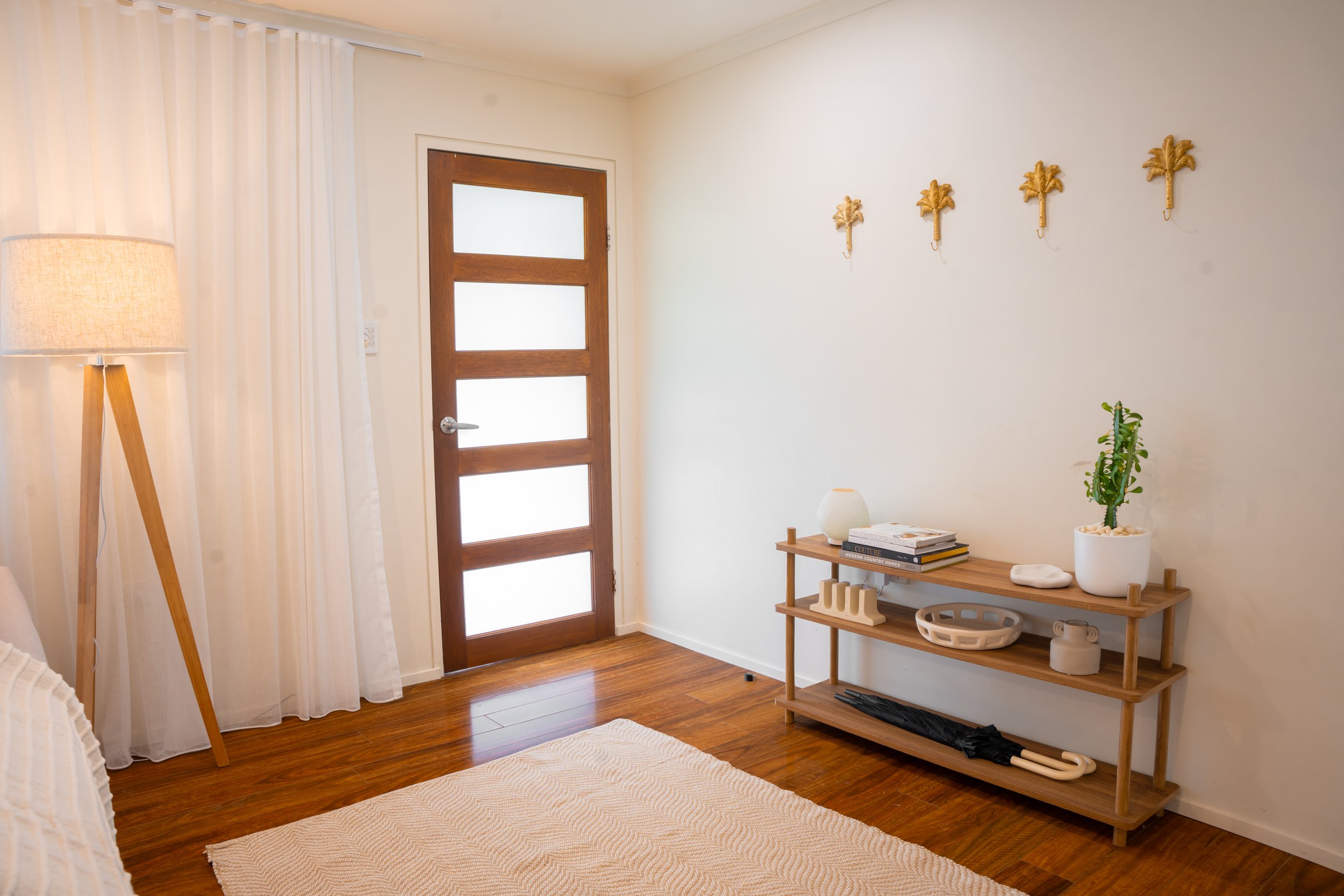 A minimalist entryway with a wooden door with frosted glass panels, a wooden console table decorated with plants, books, and decorative items, a tall wooden tripod floor lamp, white curtains, and a beige rug on polished wooden flooring.