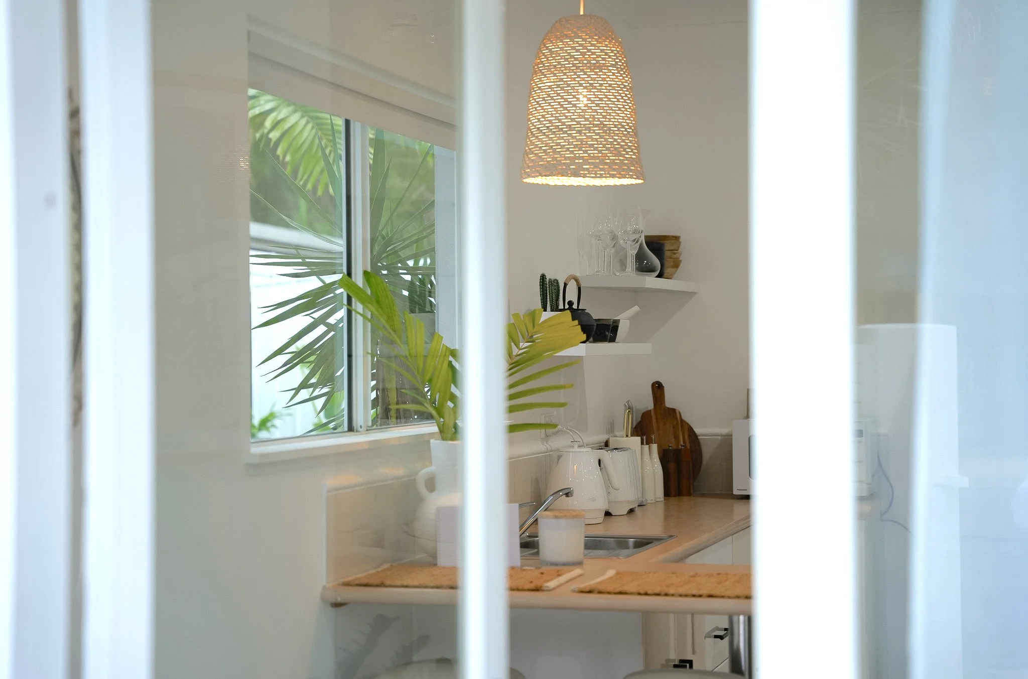 A view of a bright kitchen seen through white blinds, with a large window, hanging pendant light, shelves with glassware and plants, and a countertop with kitchen appliances.