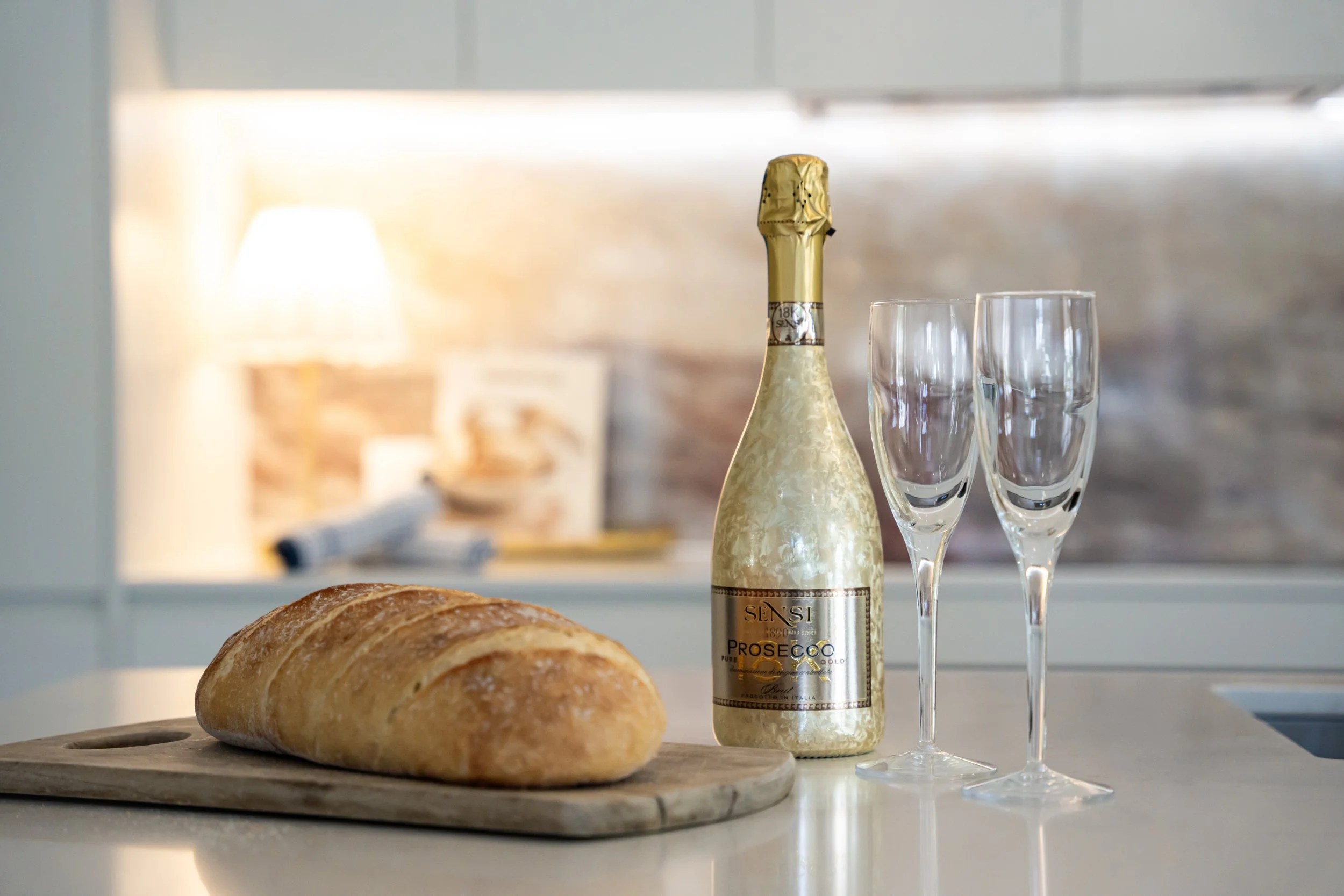 A loaf of bread on a wooden cutting board, a bottle of prosecco with a gold foil top, and two empty champagne glasses on a white countertop, with a blurred background of a kitchen.