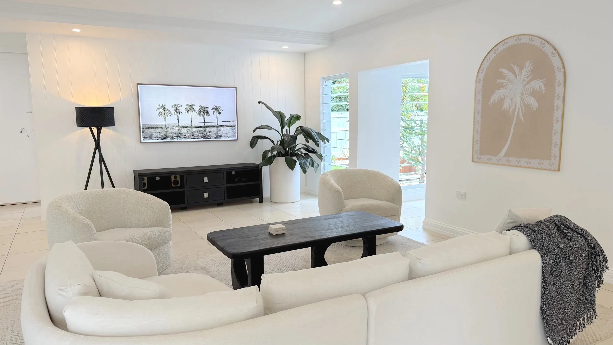 Living room with white sofa, beige armchair, black coffee table, black TV stand, wall art of palm trees, large potted plant, and windows with blinds.