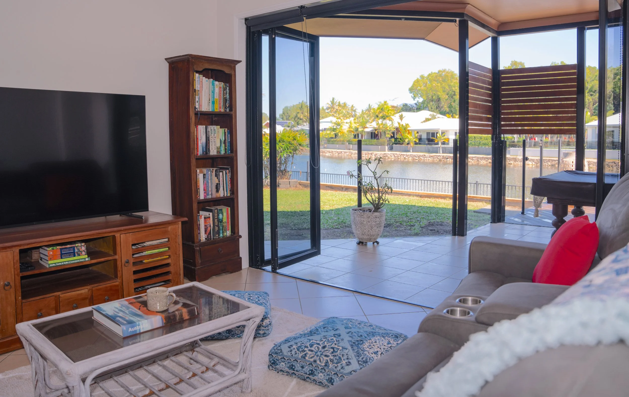 Living room with a sliding glass door leading to a backyard with water view. Includes a wooden bookshelf, a TV, a beige sofa with red and white cushions, a white coffee table with books and a cup, and decorative rugs.