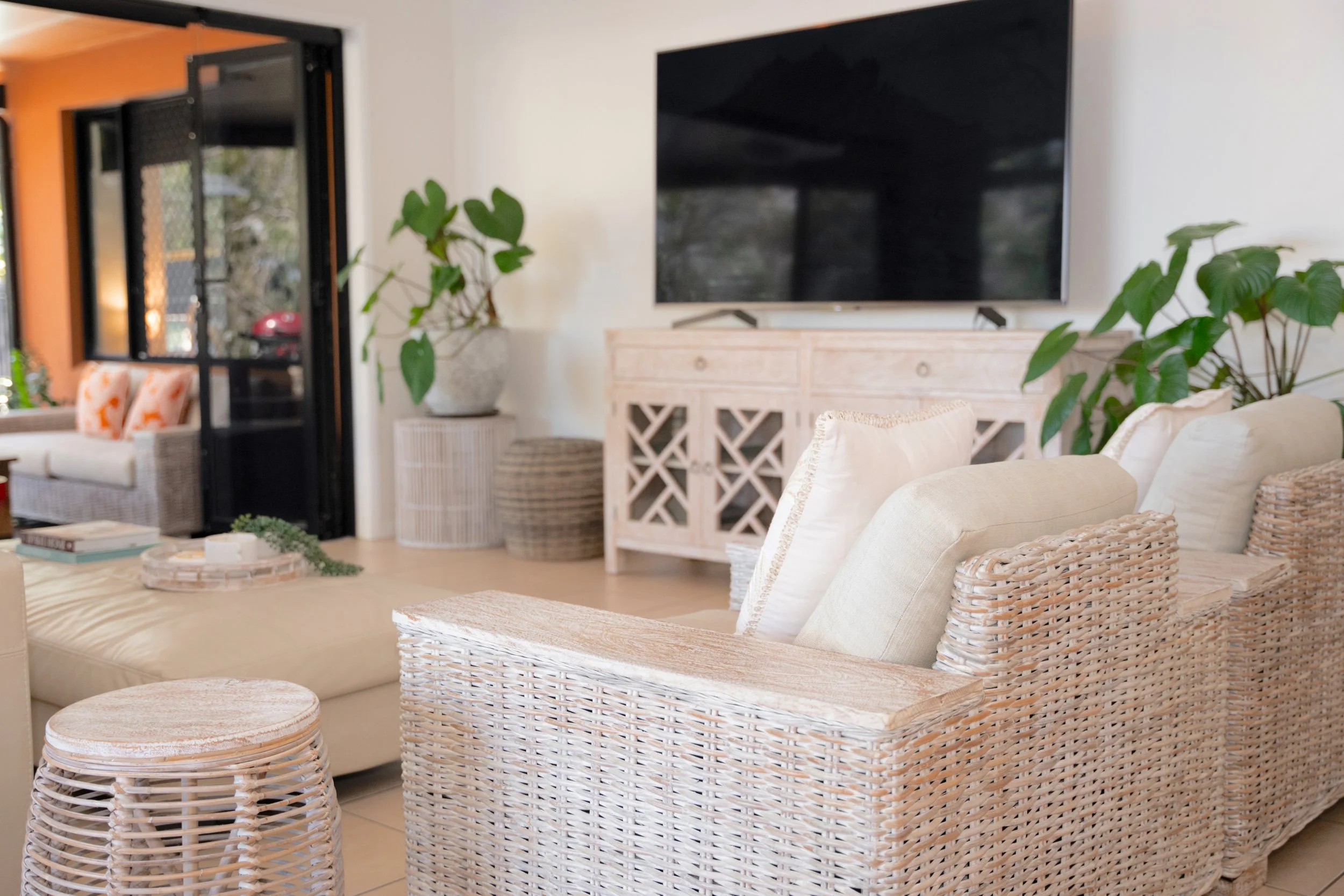 Living room with wicker chairs, a beige sofa, a wooden console with a large black television on top, and potted green plants. Part of a patio with outdoor seating is visible through glass sliding doors.