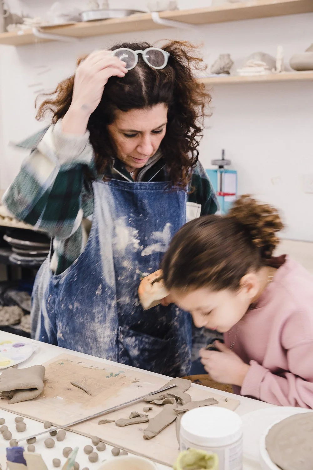 Peinture d'un vase blanc avec un motif de petits taches grises, quelqu'un en train de peindre avec un pinceau. En arrière-plan, des étagères avec des bouteilles vertes et d'autres objets.