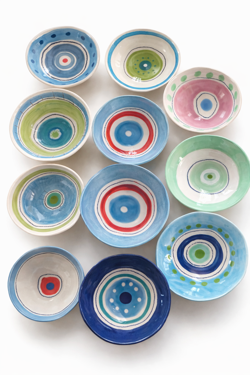 Assortiment de petites assiettes en céramique avec motifs circulaires colorés en bleu, vert, rouge, rose et blanc, disposées sur une surface blanche.