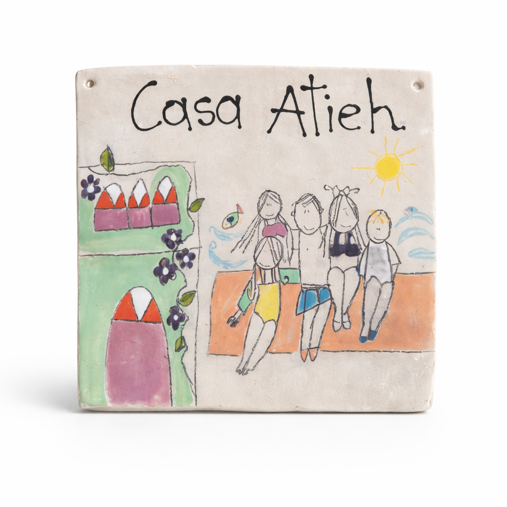 Une plaque en céramique illustrée avec le texte 'Casa Atieh', montrant quatre personnes jouant sur la plage sous un soleil, avec une maison colorée sur la côte à gauche.