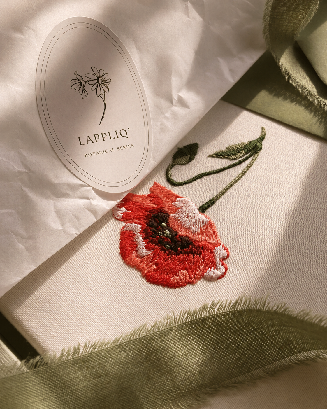 Close-up of embroidered poppy August birth flower canvas partially wrapped in glassine paper and tied with sage green ribbon.