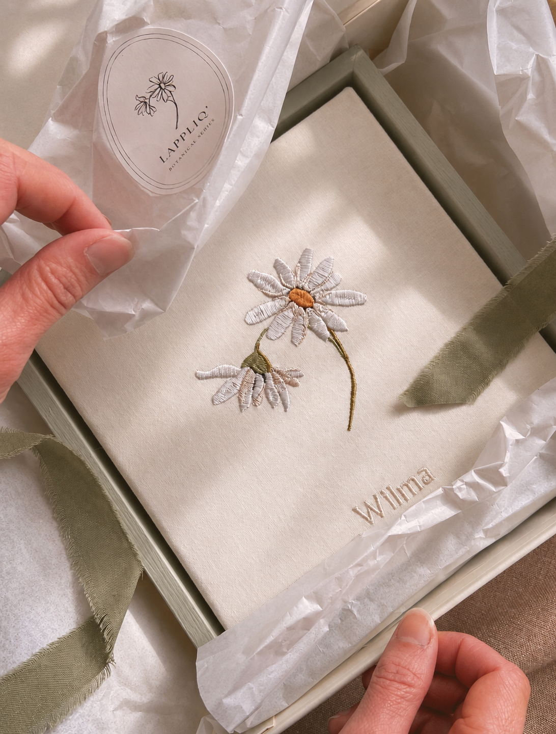 Close-up of embroidered daisy April birth flower canvas partially wrapped in glassine paper and tied with sage green ribbon.