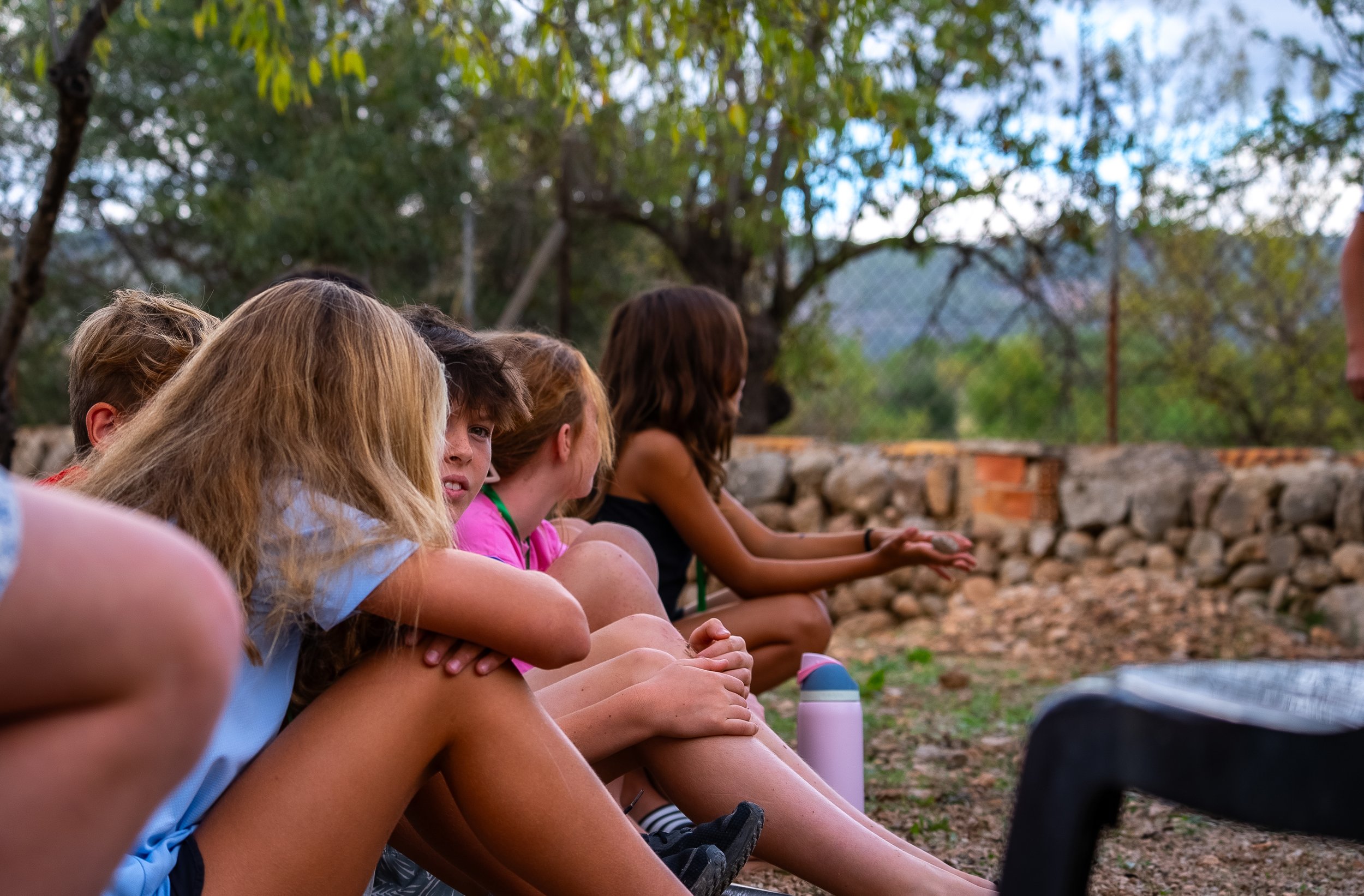 Group of children sitting outdoors on the ground during the daytime, with trees and a stone wall in the background.