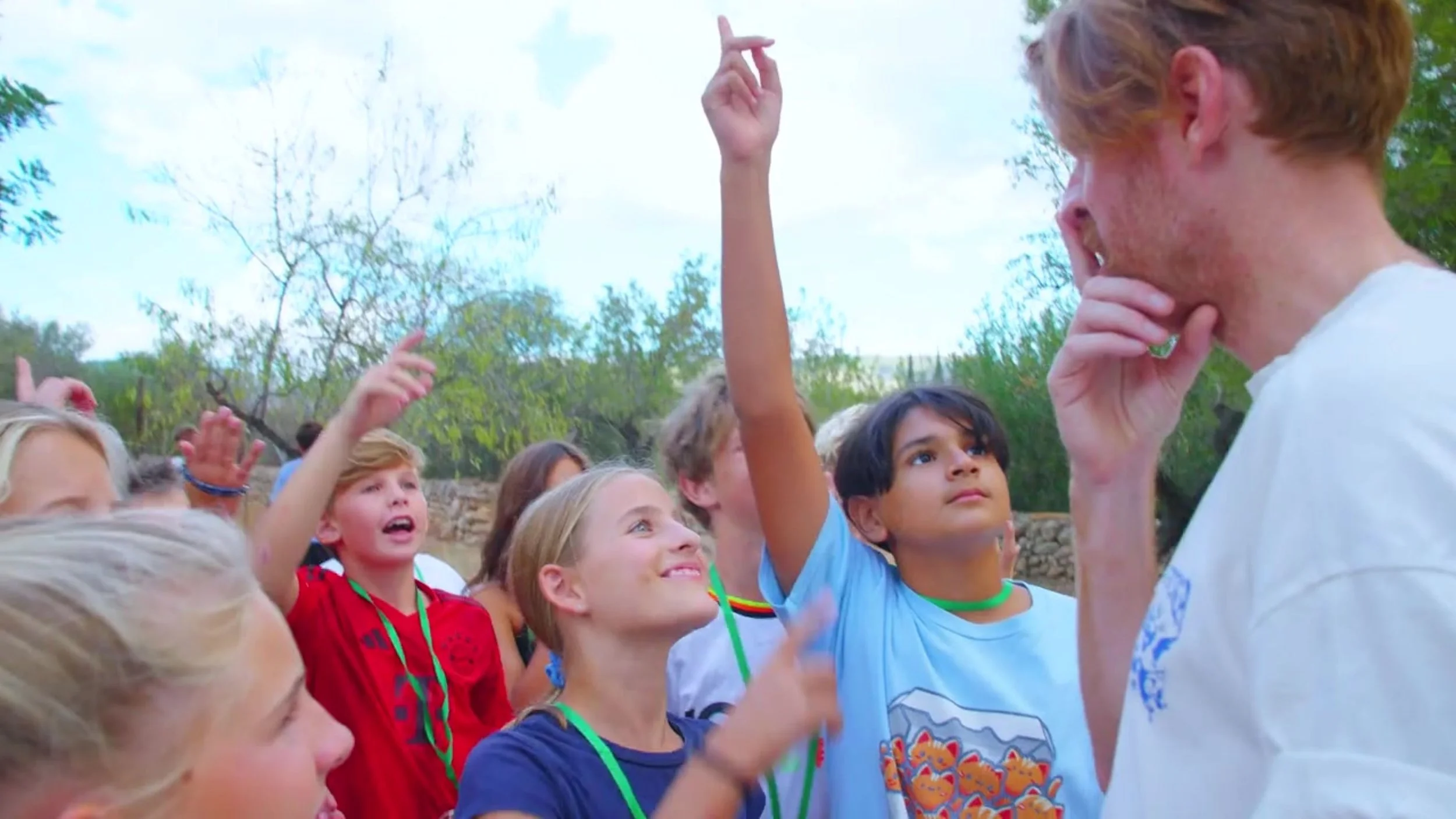 A group of children gathered outdoors, some raising their hands, listening attentively to a young man with red hair and a white shirt who appears to be speaking or teaching.