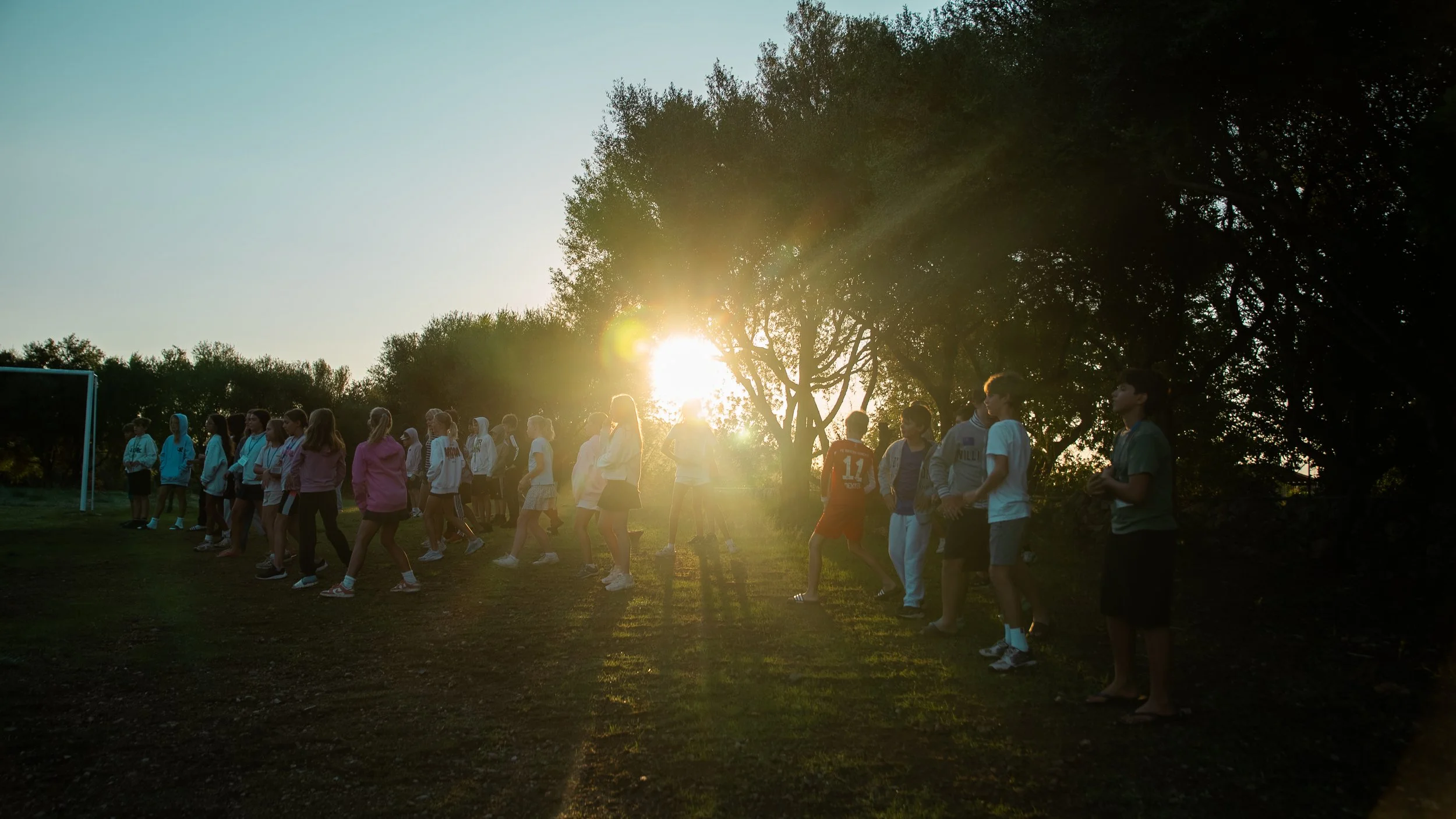 Group of children outdoors during sunset, standing in a line under trees, with sunlight shining through and casting long shadows.