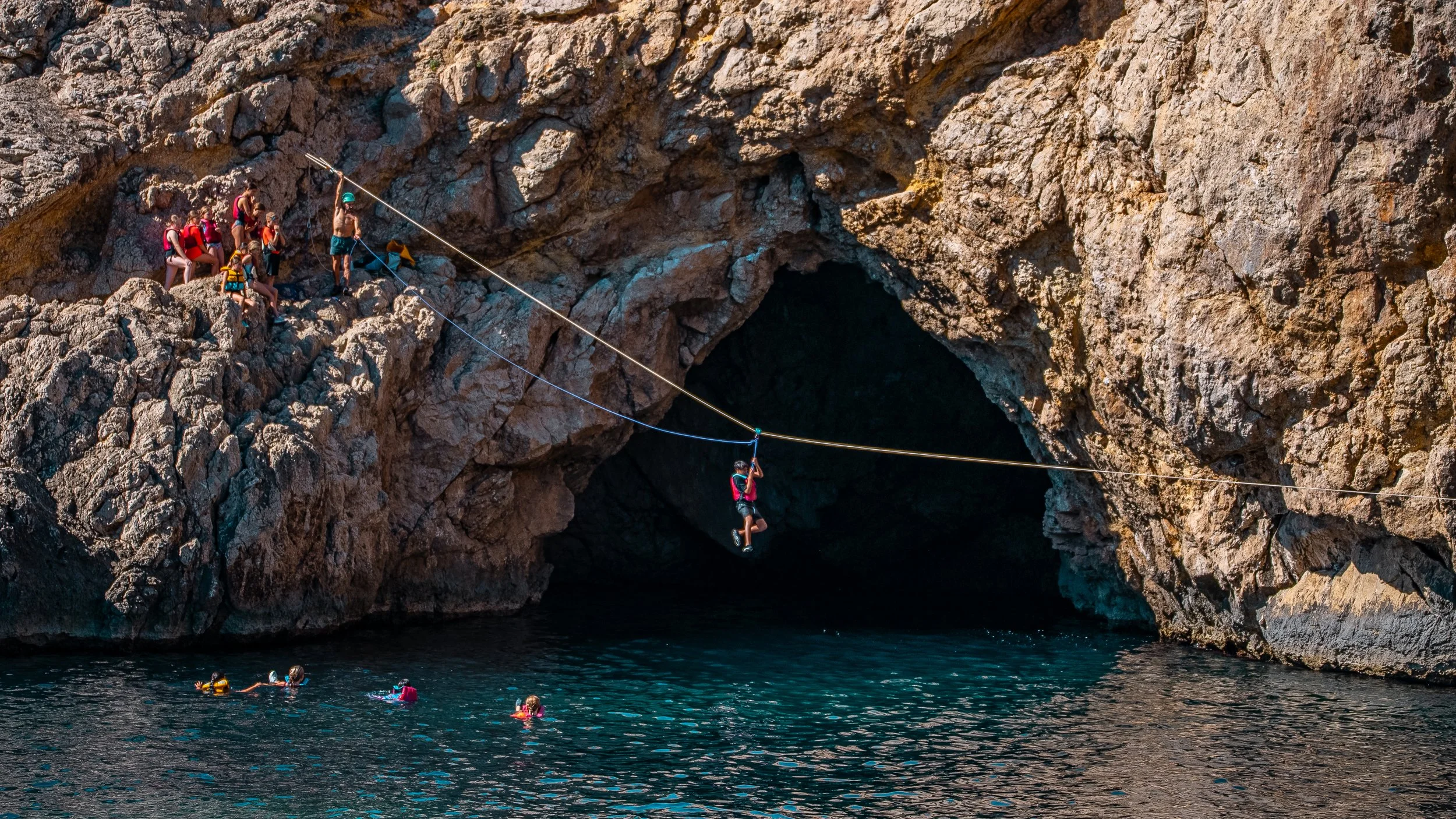 A group of people on rocky cliffs above water, with some preparing for a leap while others swim or wait in the water, near a large cave entrance.