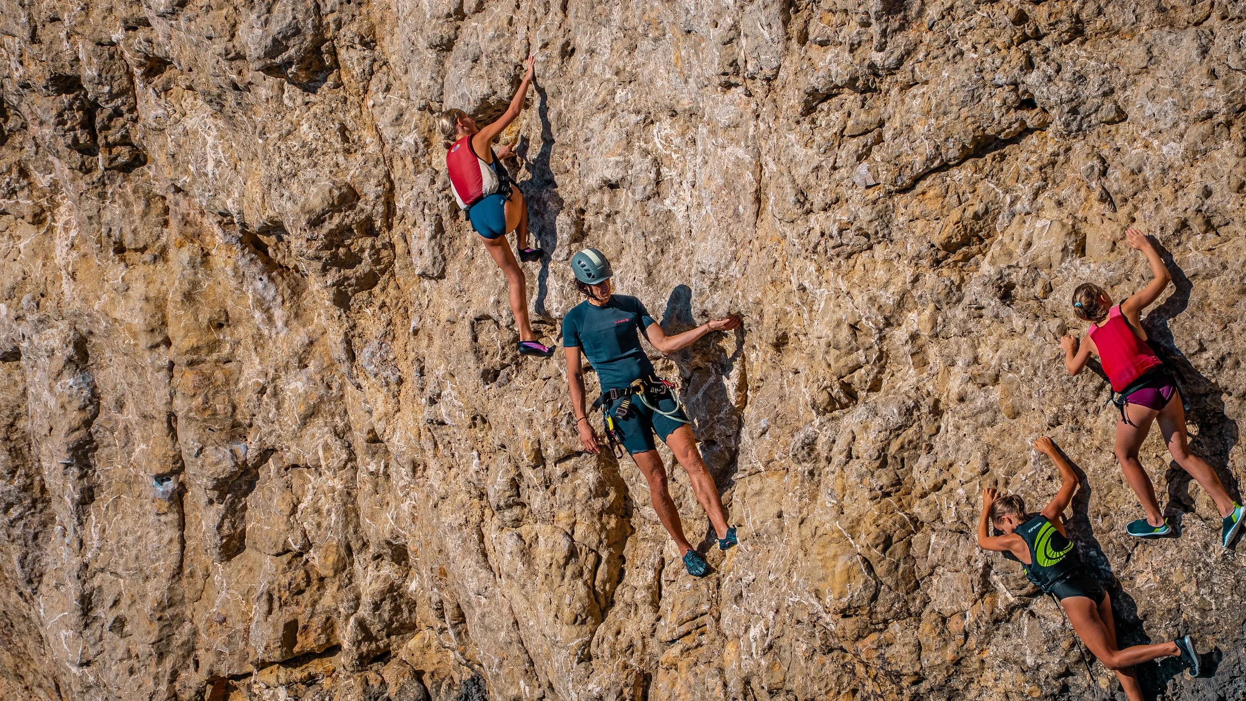 Four people rock climbing on a steep outdoor cliff, wearing helmets and harnesses, with two on the left reaching higher and two on the right climbing downward.
