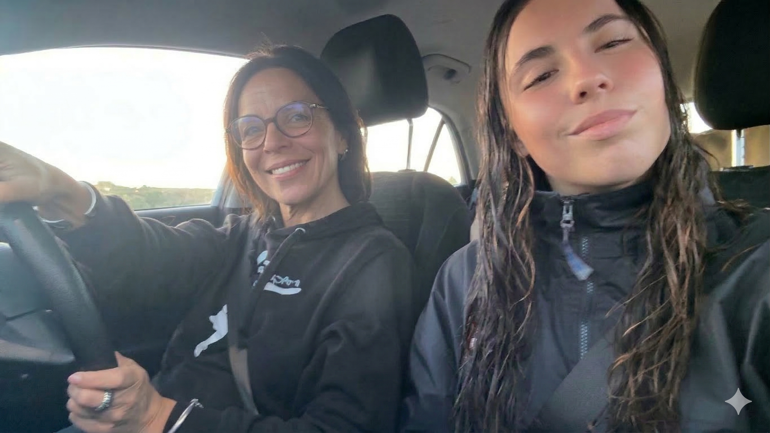 Two women sitting in a car, smiling at the camera. The woman on the left has glasses and short dark hair, wearing a black hoodie. The woman on the right has long, wet, wavy hair, and is wearing a black jacket, with seat belts fastened.