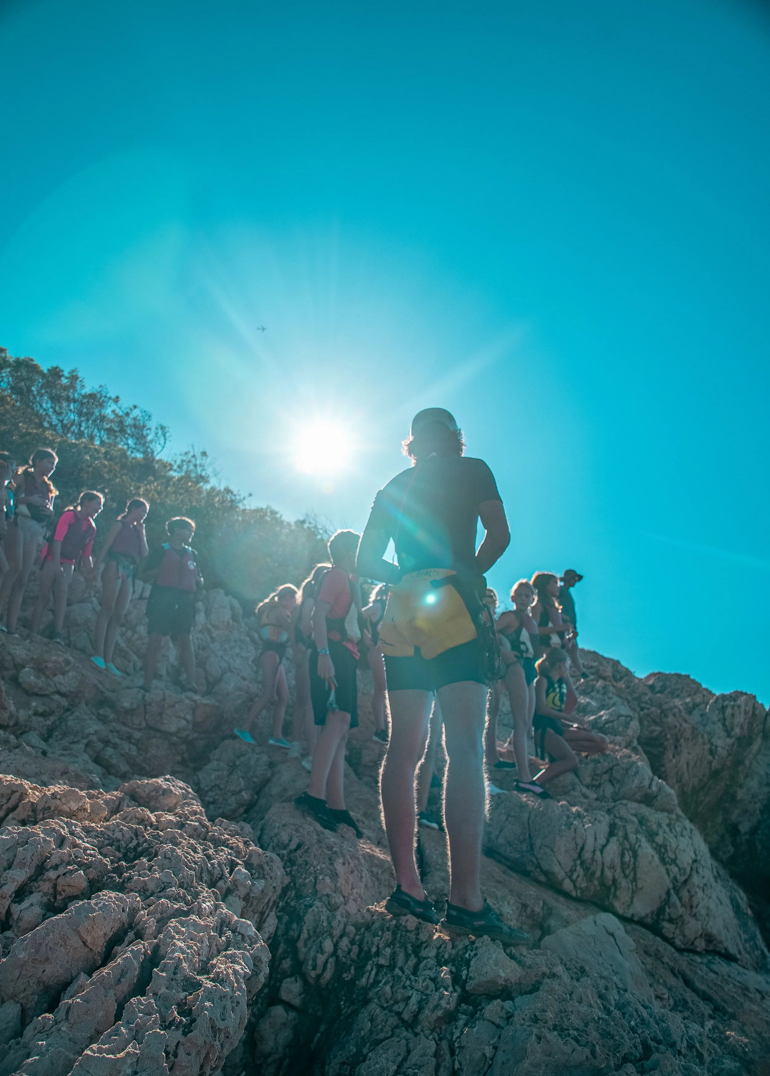 A group of people hiking on rocky terrain under a bright blue sky, with the sun shining brightly in the background.