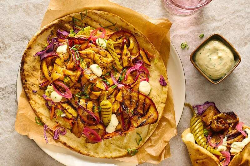 Flatbread belegd met gegrilde gekruide auberginereepjes, rode kool, ingelegde rode ui, lente-ui en witte sausdruppels, met een gevouwen wrap en kommetje saus ernaast