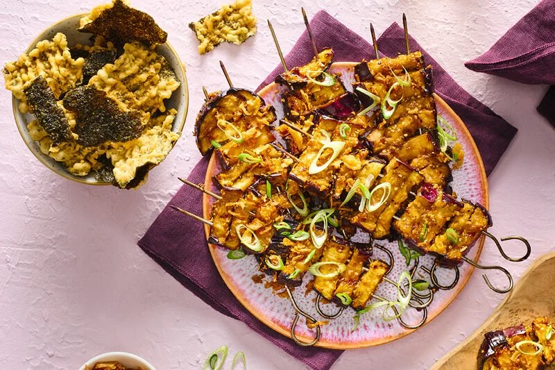 Gegrilde aubergineblokjes op spiesen met goudbruin misoglazuur en lente-ui op een roze bord, naast een kom met nori zeewier chips