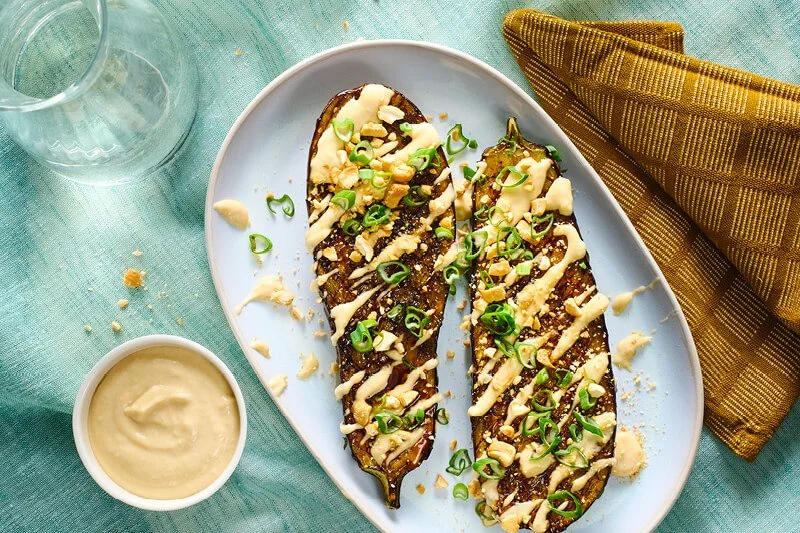 Twee helften gegrilde aubergine met donkere teriyakiglazuur, besprenkeld met romige gele saus, gehakte noten en gesneden lente-ui op een wit ovaal bord, met een kommetje saus erbij