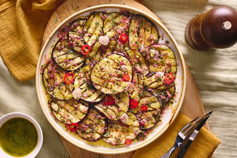 Dunne gegrilde aubergineschijven cirkelvormig uitgewaaierd op een groot bord, overgoten met groene kruidenolie, rode chiliplakjes en fijngesneden rode ui