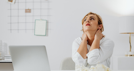 women dressed in white sitting at a computer with her hands on the back of her neck signaling tension and stress