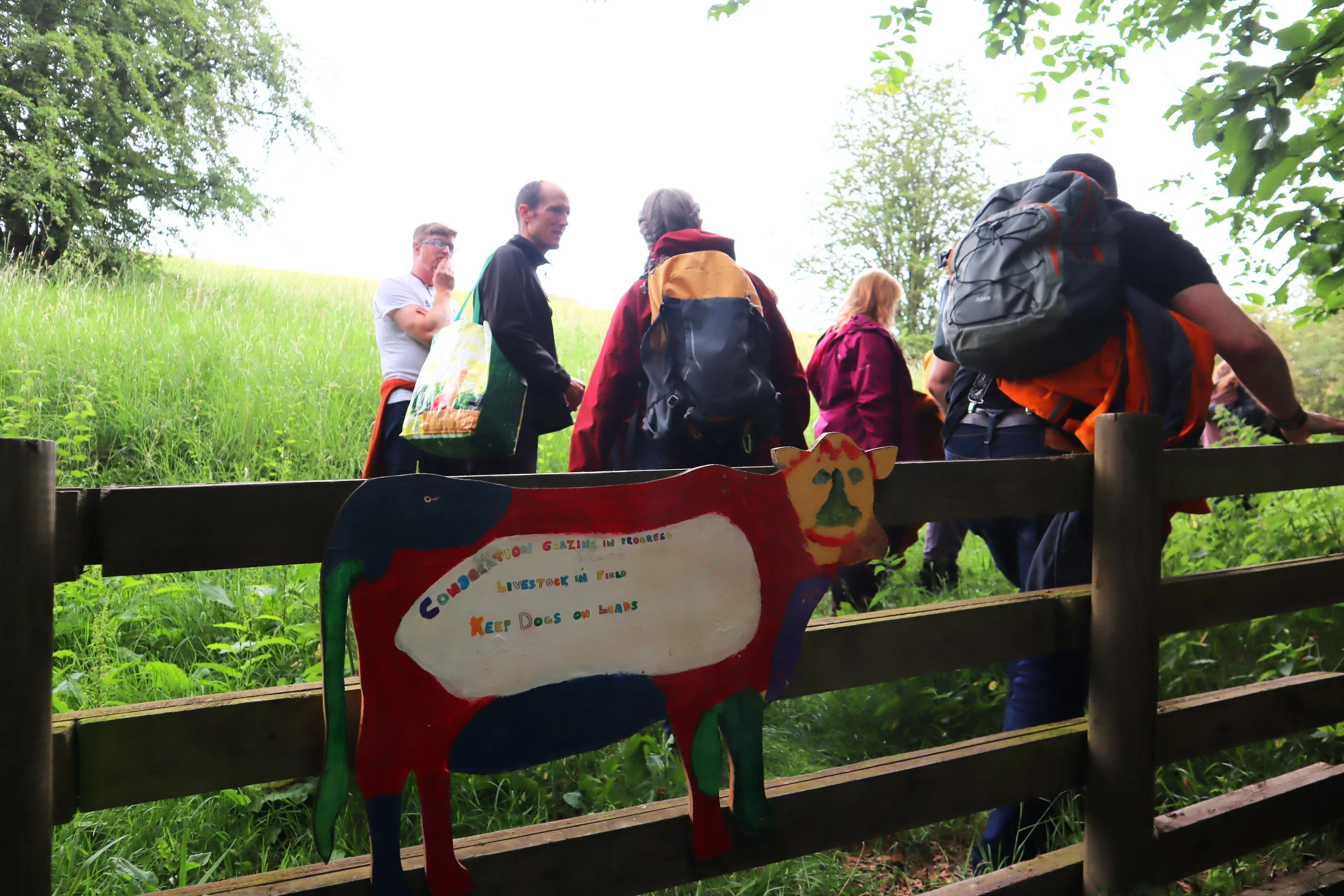 A group of walkers climb a fence. Attached to the fence is a sign shaped like a cow which reads: conservation grazing in progress, livestock in field, keep dog on lead.