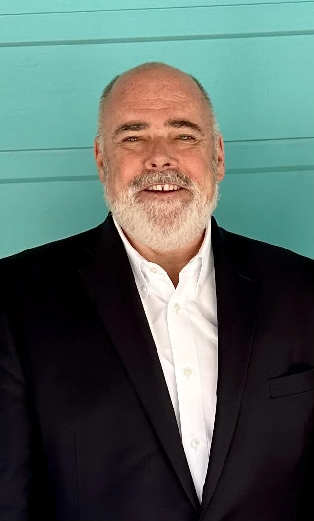 A smiling middle-aged man with a beard, dressed in a black suit and white shirt, standing in front of a teal wall.