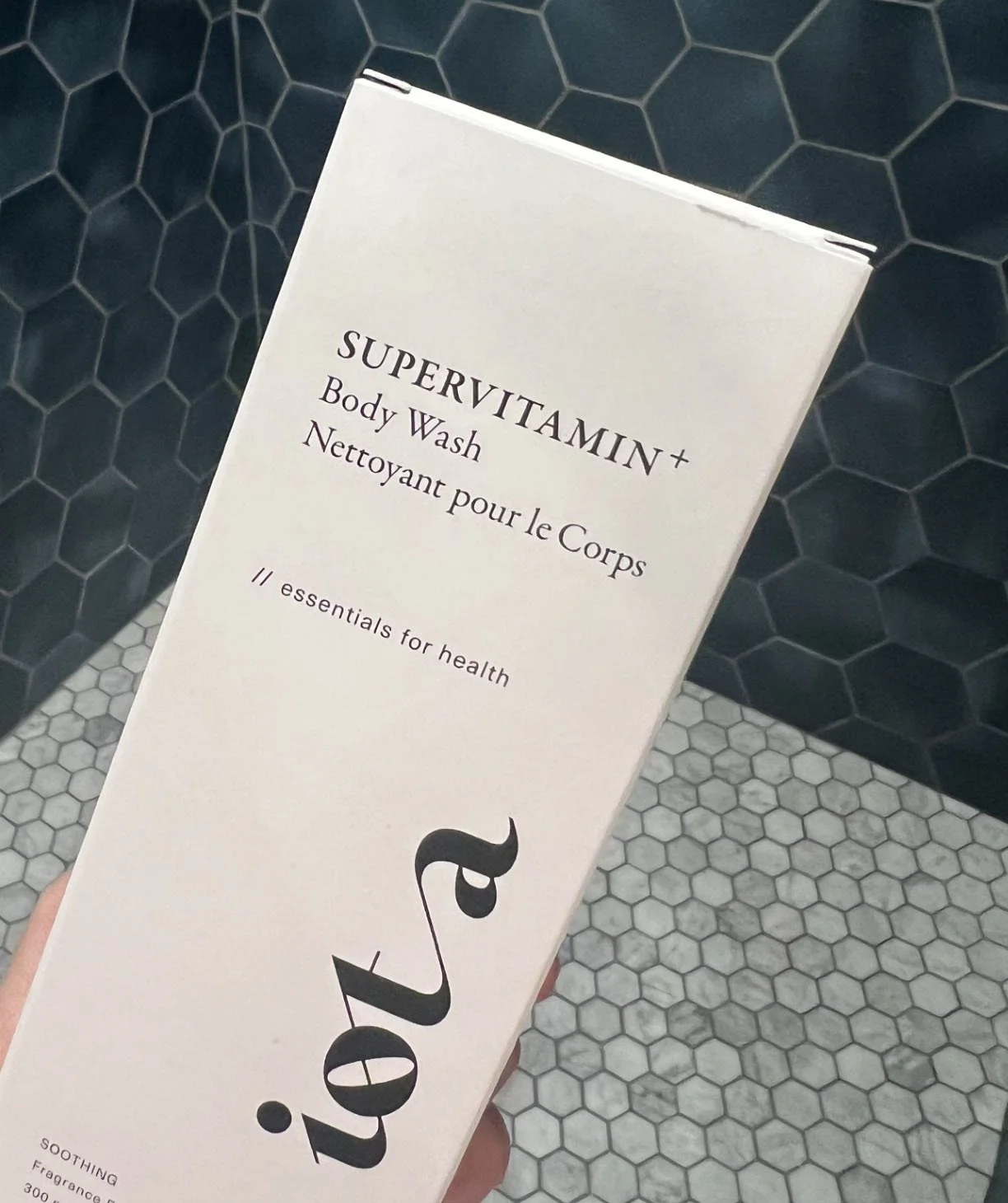 A box of Supervitamin+ Body Wash held over a tiled floor with black hexagonal tiles and smaller gray hexagonal tiles.