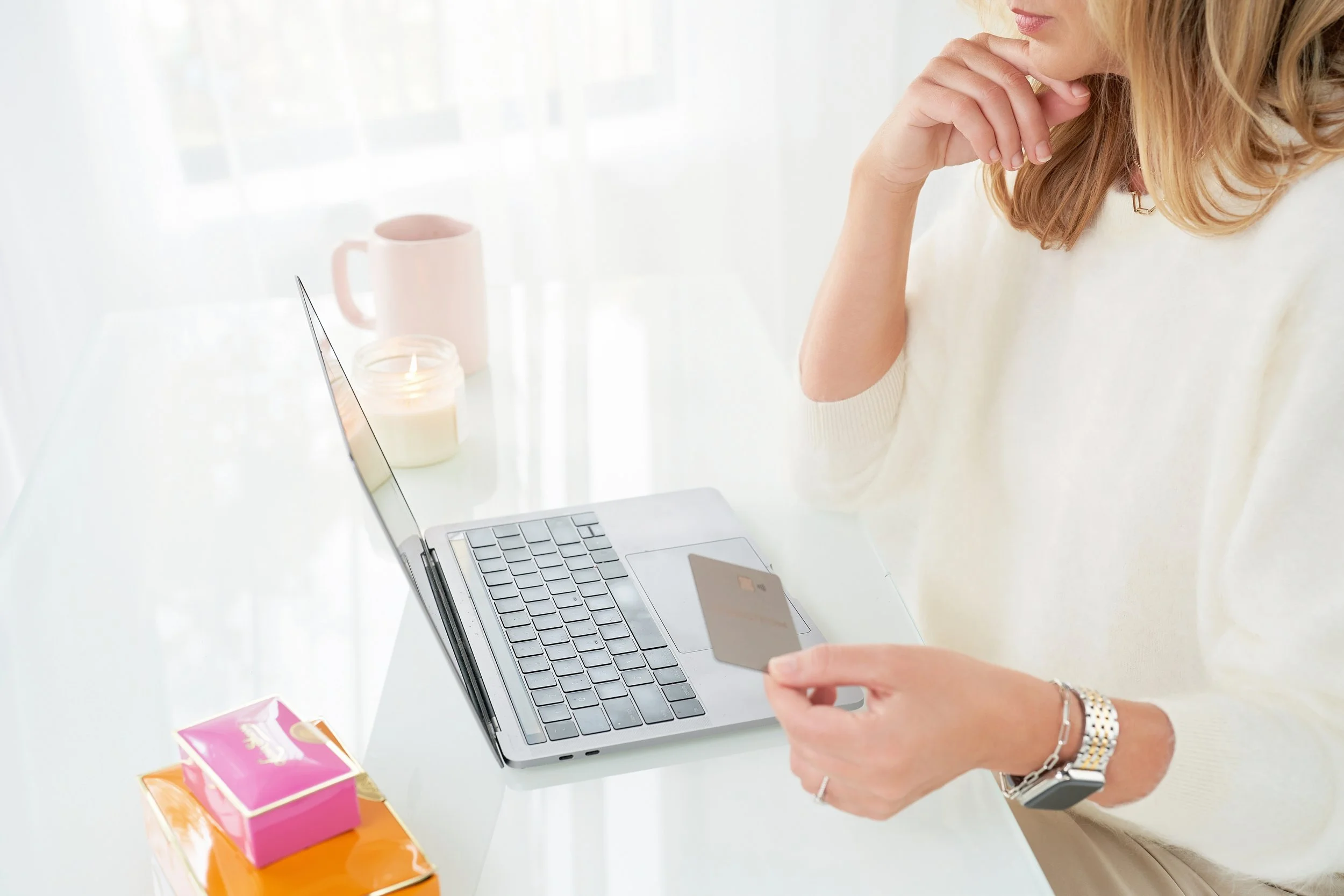 Woman shopping online on laptop, holding a credit card, with a pink mug, candle, and tissue box on white desk.