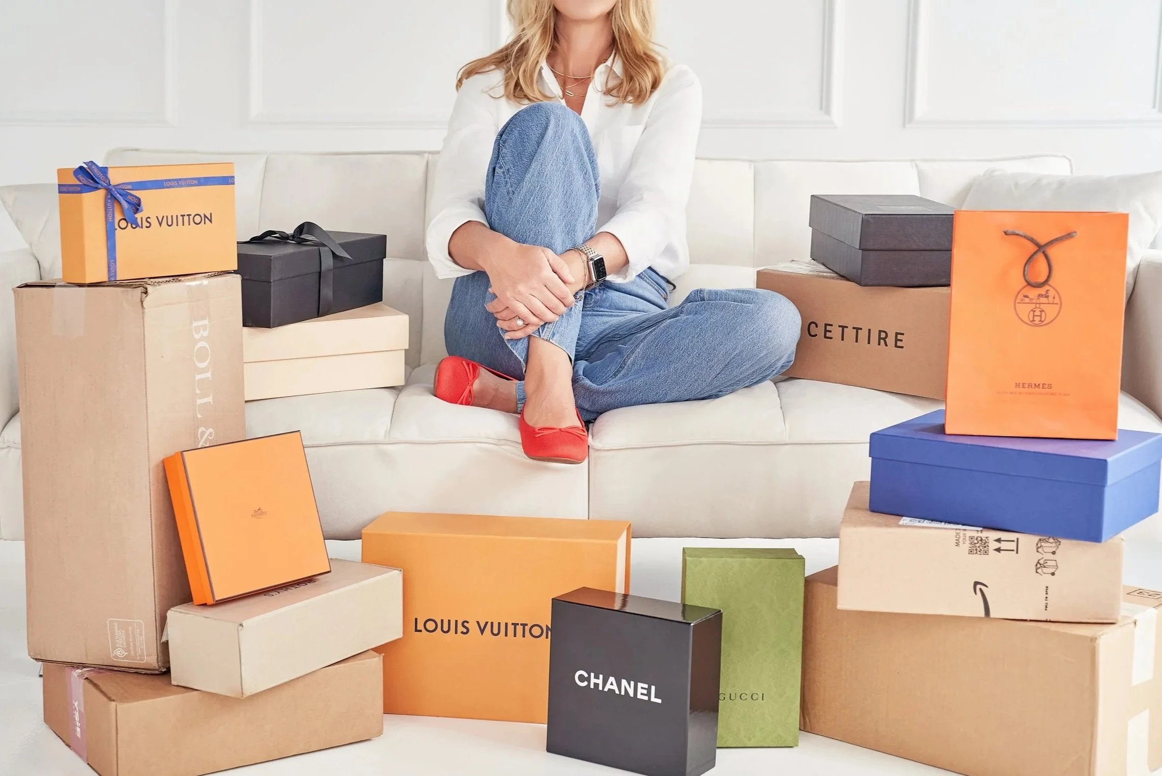 A woman sitting on a white sofa surrounded by numerous shopping boxes from luxury brands like Louis Vuitton, Chanel, Gucci, Hermès, and Cettire.