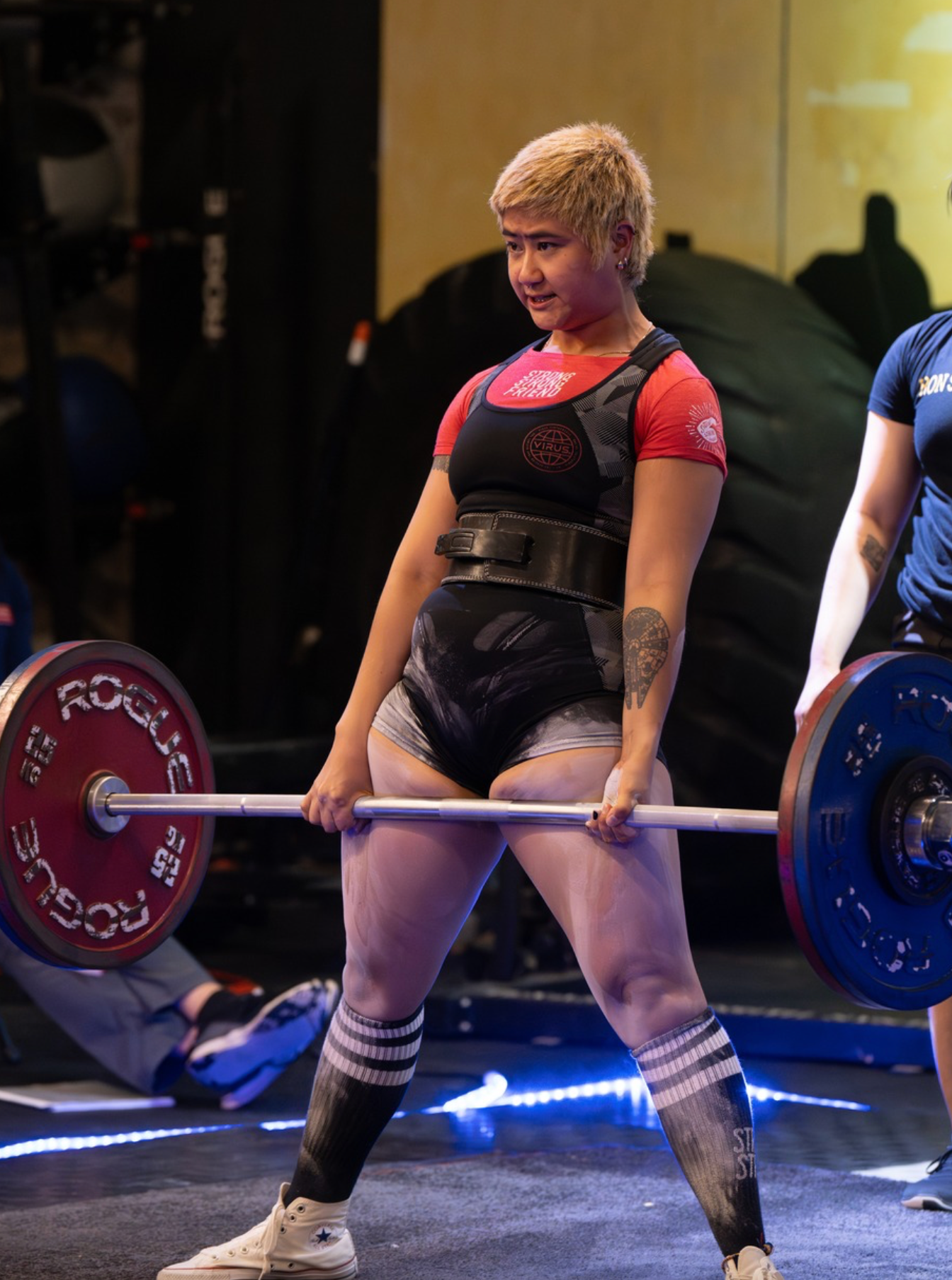 A woman with short blonde hair and tattoos on her arms, wearing a red shirt and black shorts, is lifting a barbell during a weightlifting session in a gym.