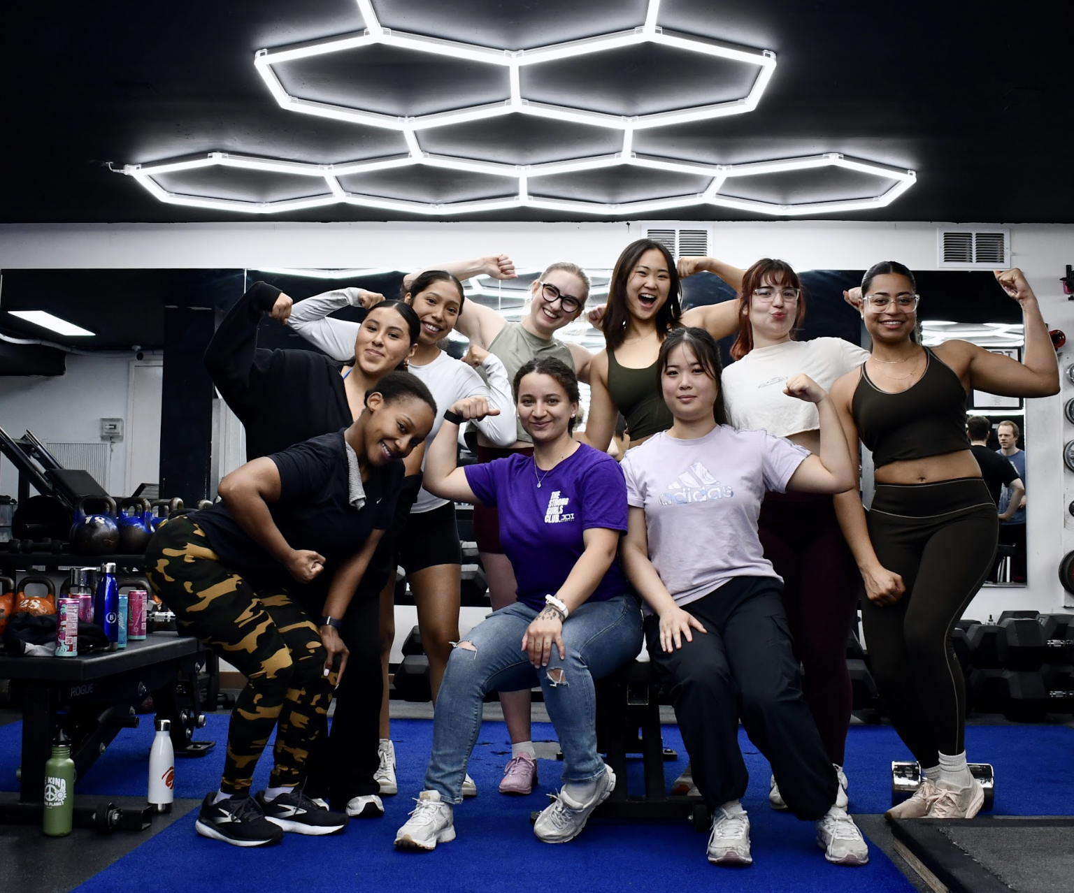 Group of women in a gym showing their arm muscles, smiling and flexing, with weight equipment and water bottles in the background.