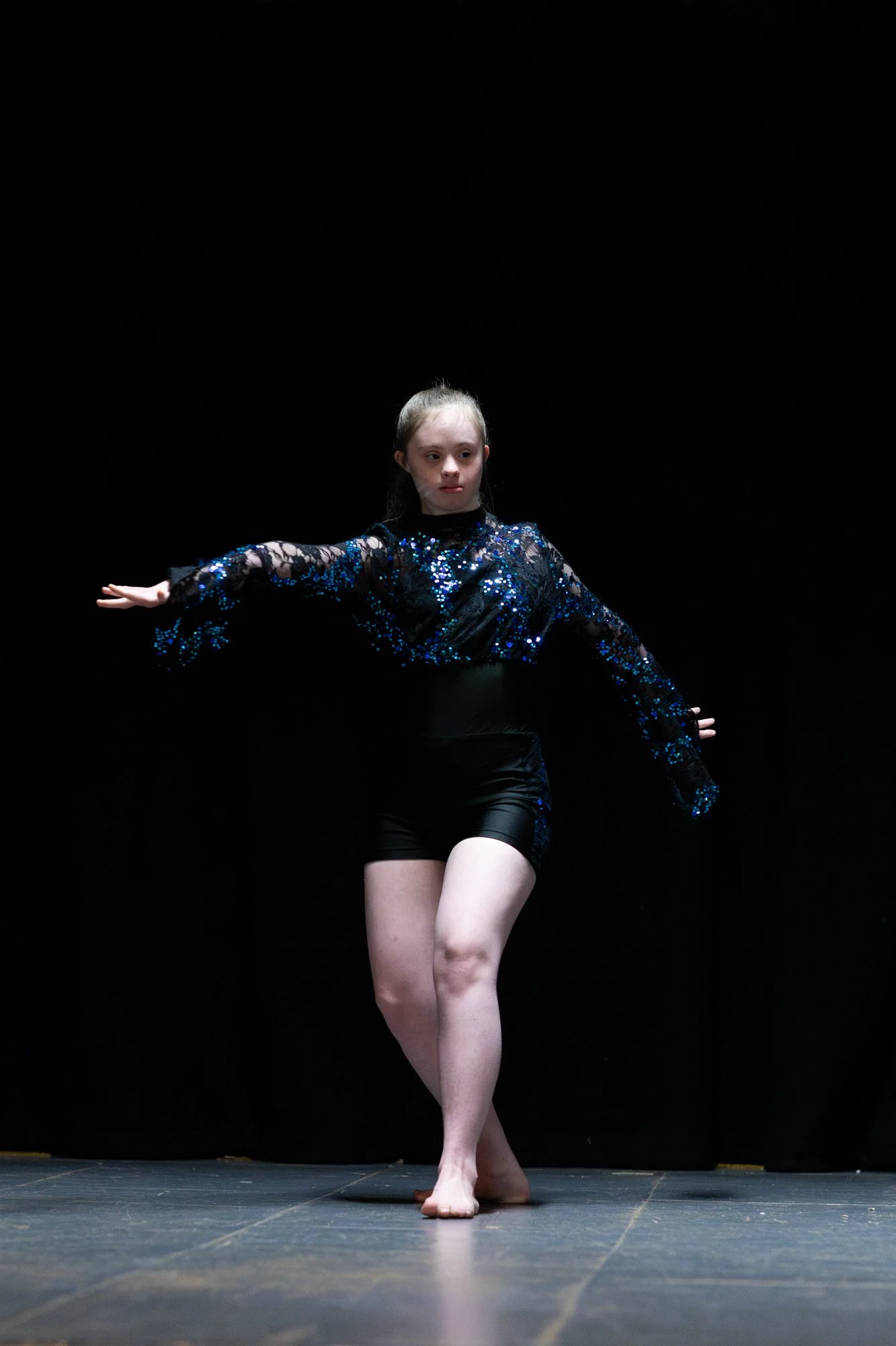 A young girl performing a dance on stage with a black background, wearing a black and blue sequined costume.