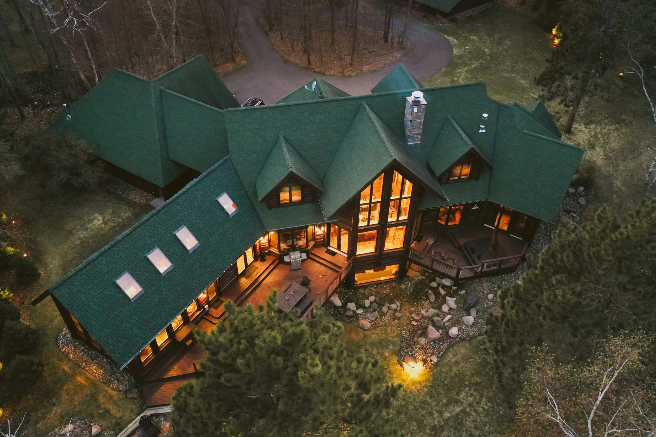 Aerial view of a large house with a green roof, illuminated from inside, surrounded by trees and a yard at dusk.