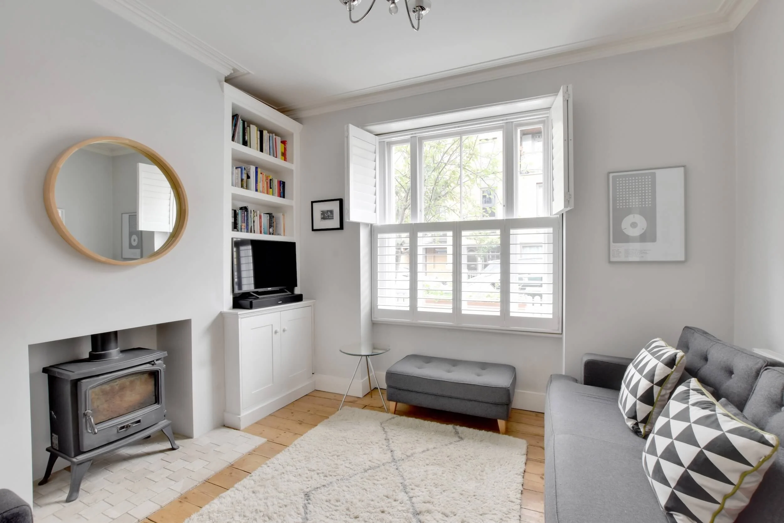 Living room with white walls, hardwood floors, a window with shutters, a gray sofa with black, white, and green patterned pillows, a small glass side table, dark gray ottoman, built-in shelving with books and a flat-screen TV, a white fireplace with 