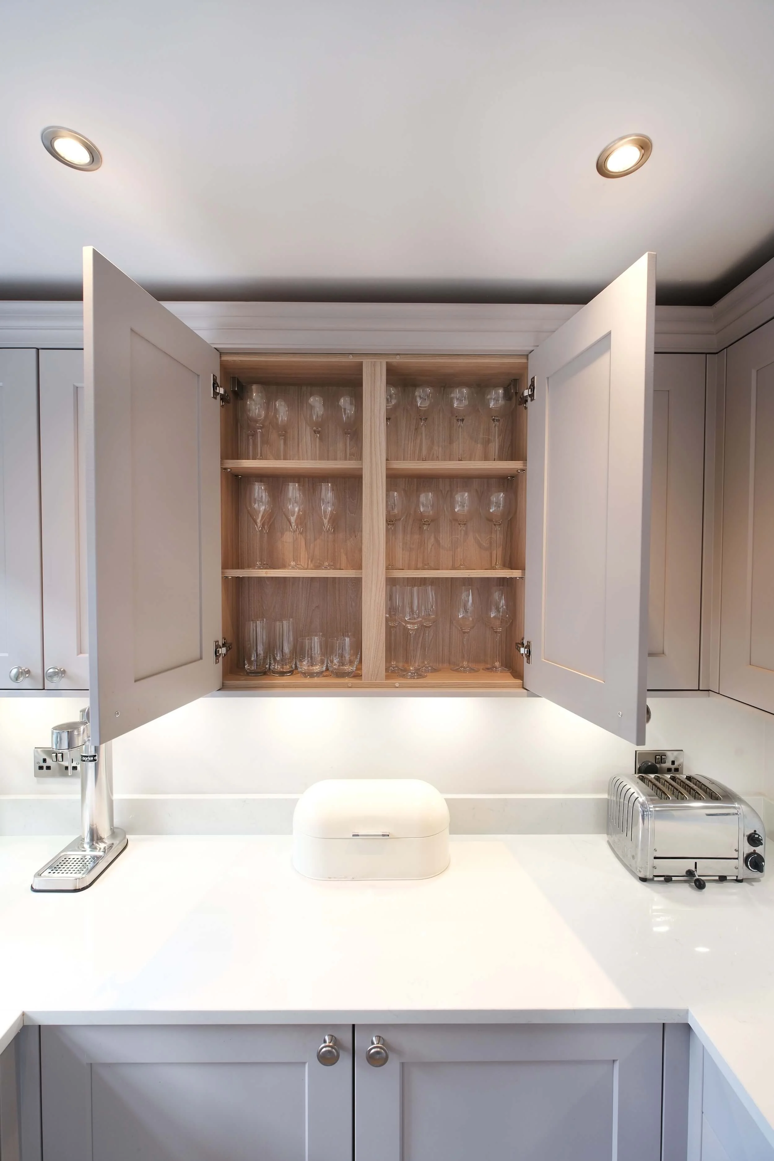 Open kitchen cabinet with shelves holding various glassware, including champagne flutes, wine glasses, and tumblers, above a white countertop with a toaster and a bread box.