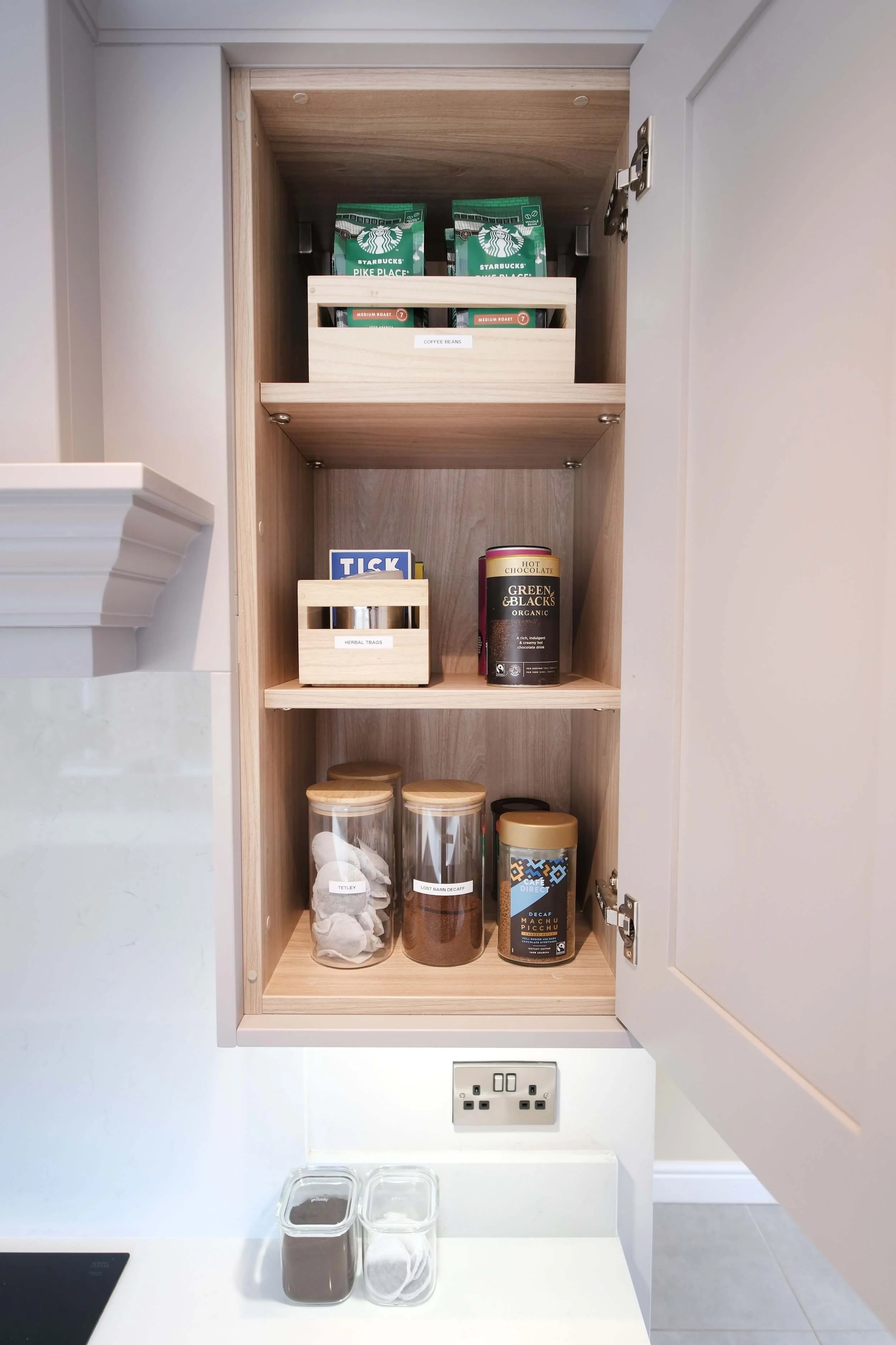 Interior view of a kitchen cabinet with shelves holding coffee and tea items, including coffee bags, tea boxes, and jars of tea leaves and coffee, with an electrical outlet and storage containers below.