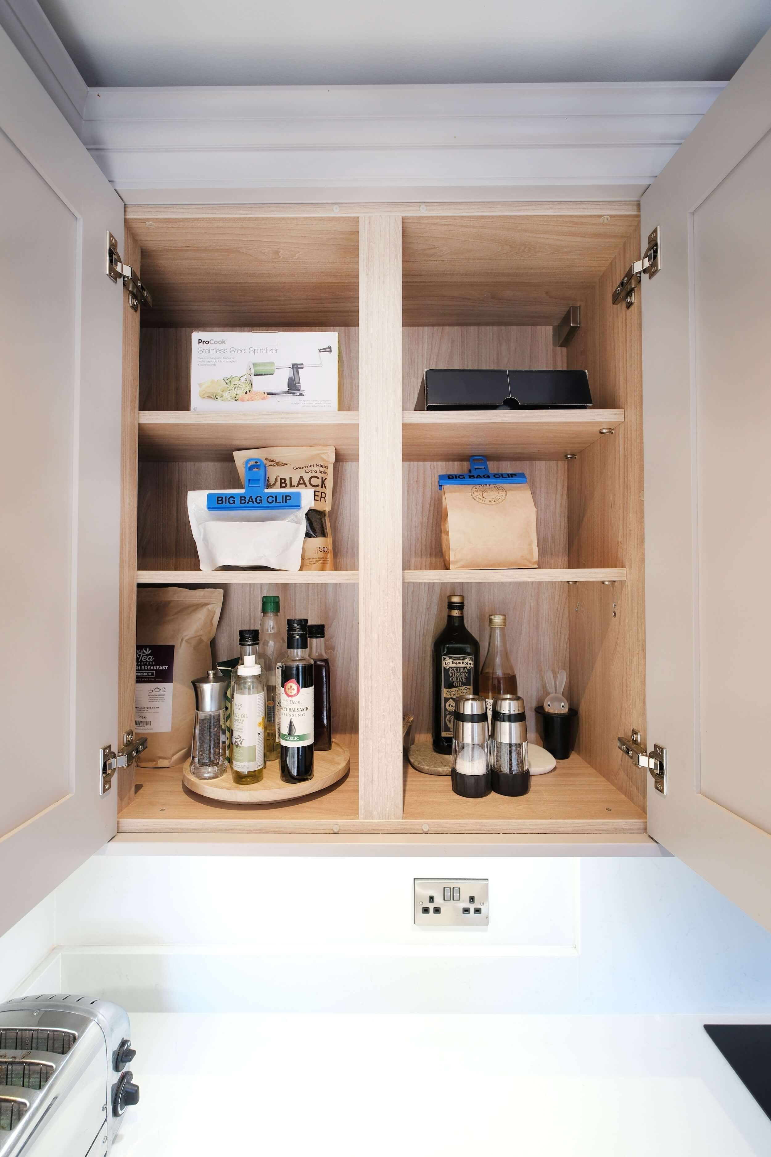 An open wooden kitchen cabinet with various food and seasoning items, a black box, and a white electrical outlet below.