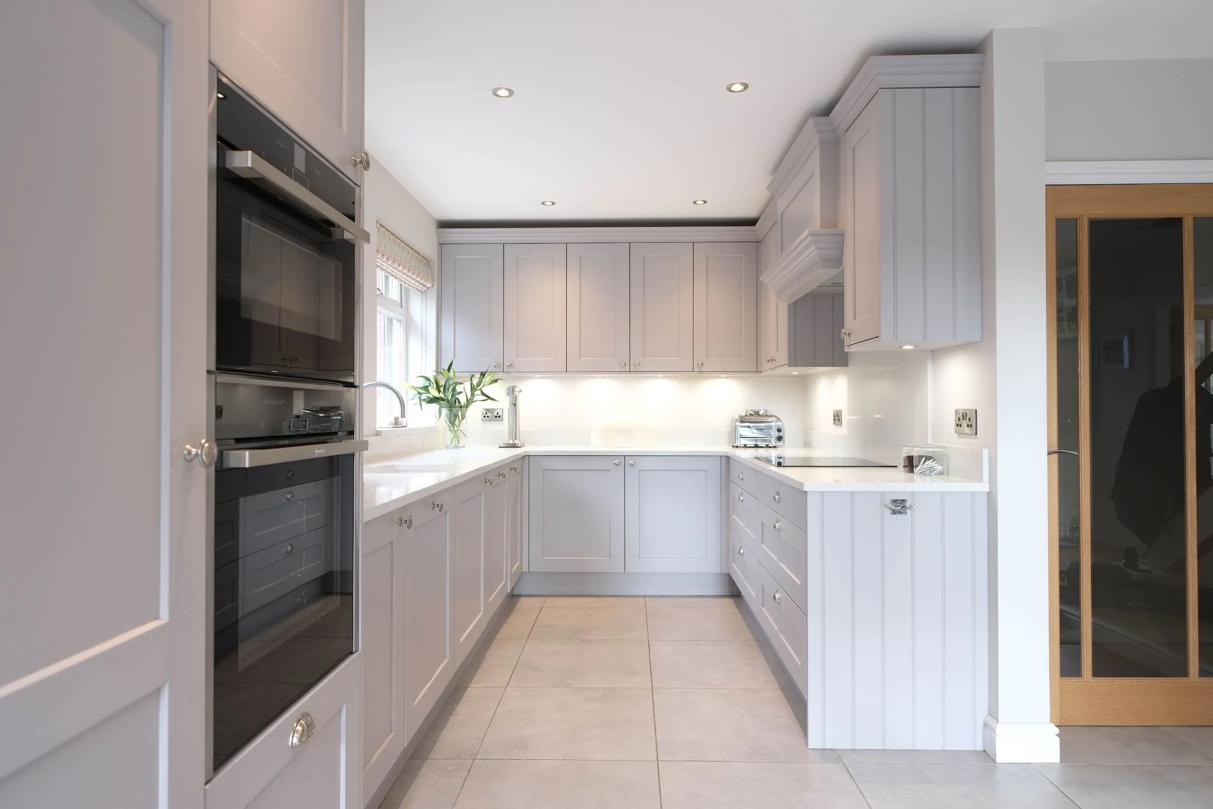 Bright, modern kitchen with white cabinetry, beige tiled flooring, a window, a stainless steel oven and microwave, a small potted plant near the sink, and a stainless toaster.