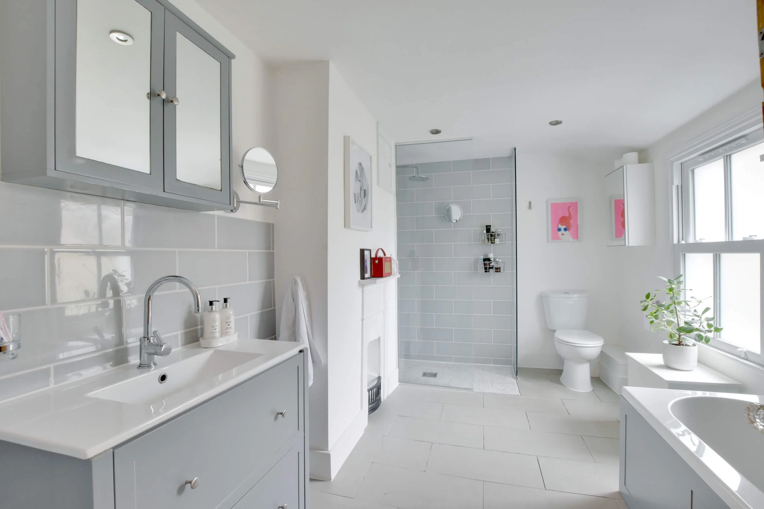 Bright, modern bathroom with grey and white color scheme, featuring a sink with a mirror cabinet, a walk-in shower with grey tiles, a toilet, large windows with a potted plant, and pink artwork on the walls.