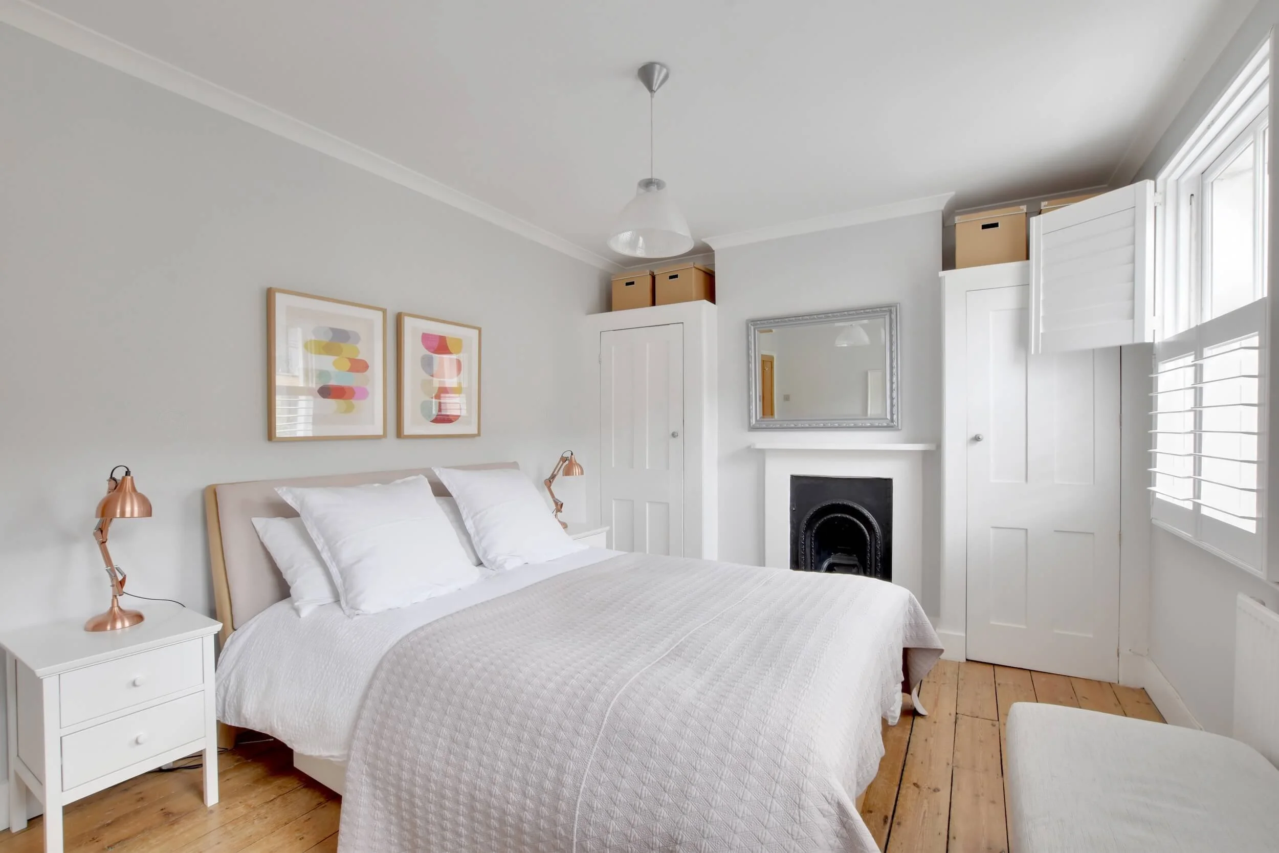 A bedroom with white walls, a white bed with pillows, a white nightstand with a copper desk lamp, above the bed are two colorful abstract art pieces, a window with white shutters, a mirror above the fireplace, and storage boxes on top of white cabine