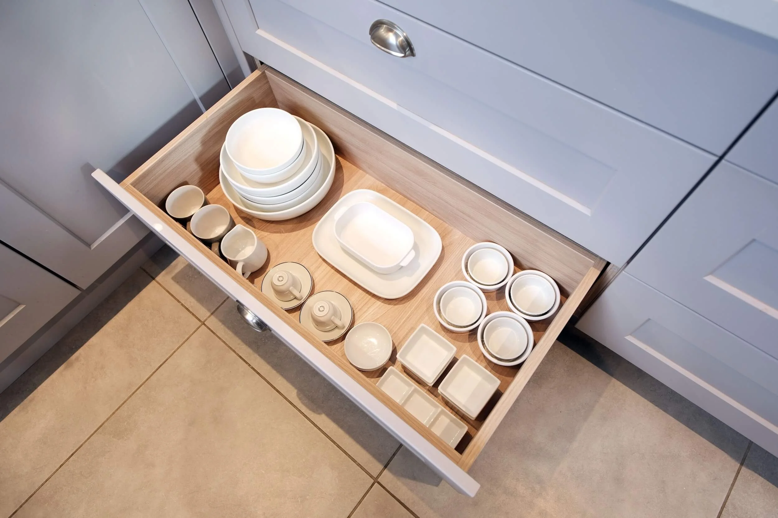 Open kitchen drawer containing white dishes, bowls, and cups on a wooden tray.