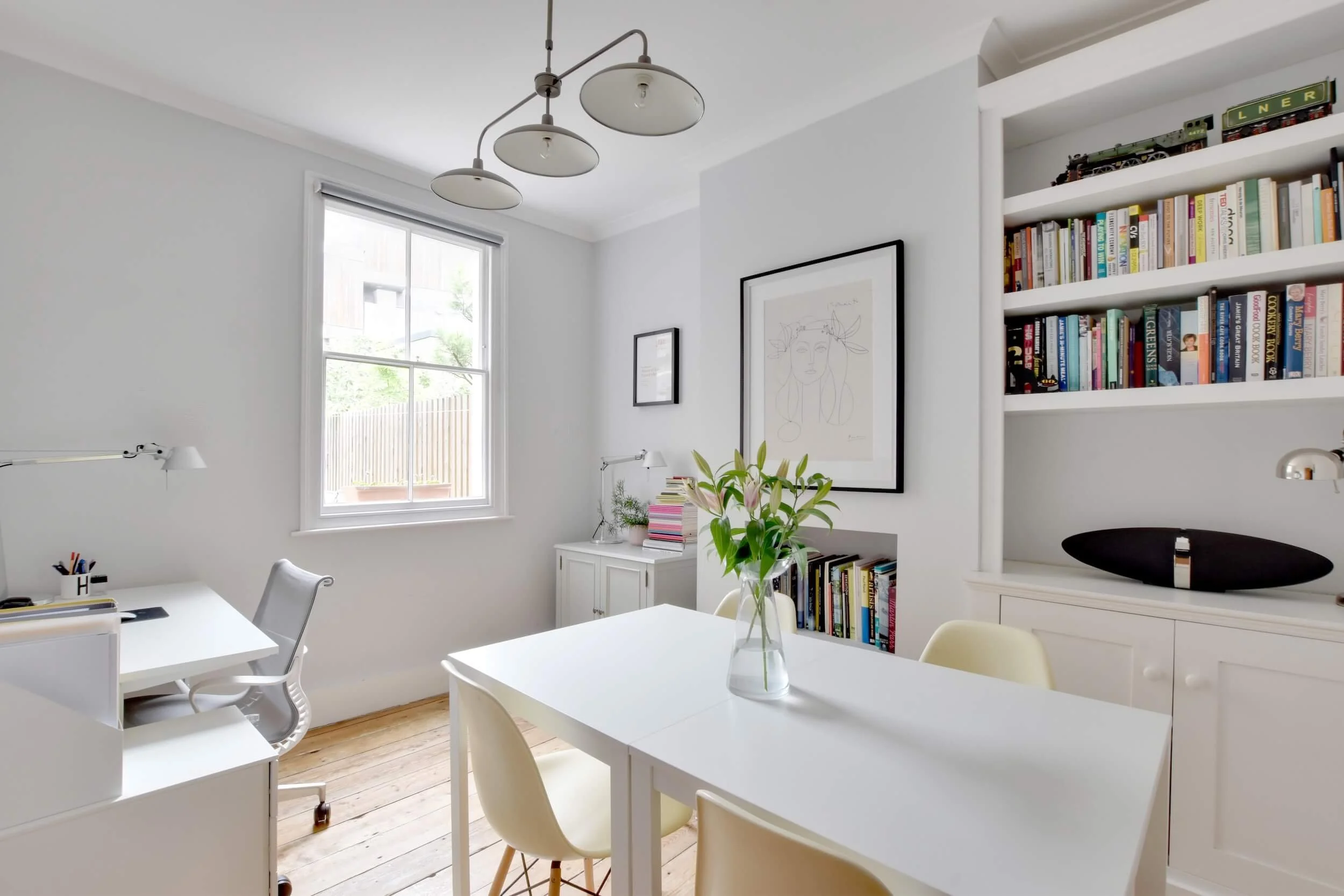 Bright, minimalistic home office with white walls, wooden floor, a window, a white desk with office chair, a white dining table with chairs, a bookshelf, and artwork on the walls.