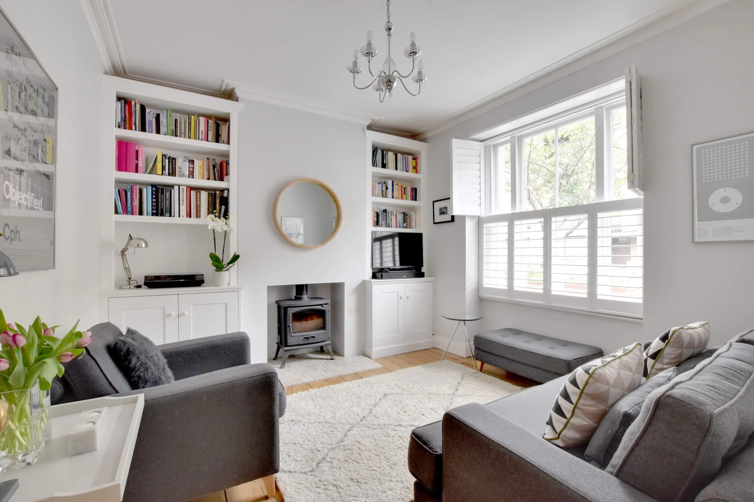 Bright living room with white walls, large window with white shutters, grey sofas, a white fluffy rug, a bookshelf with neat books, a fireplace, a TV, and pillows.